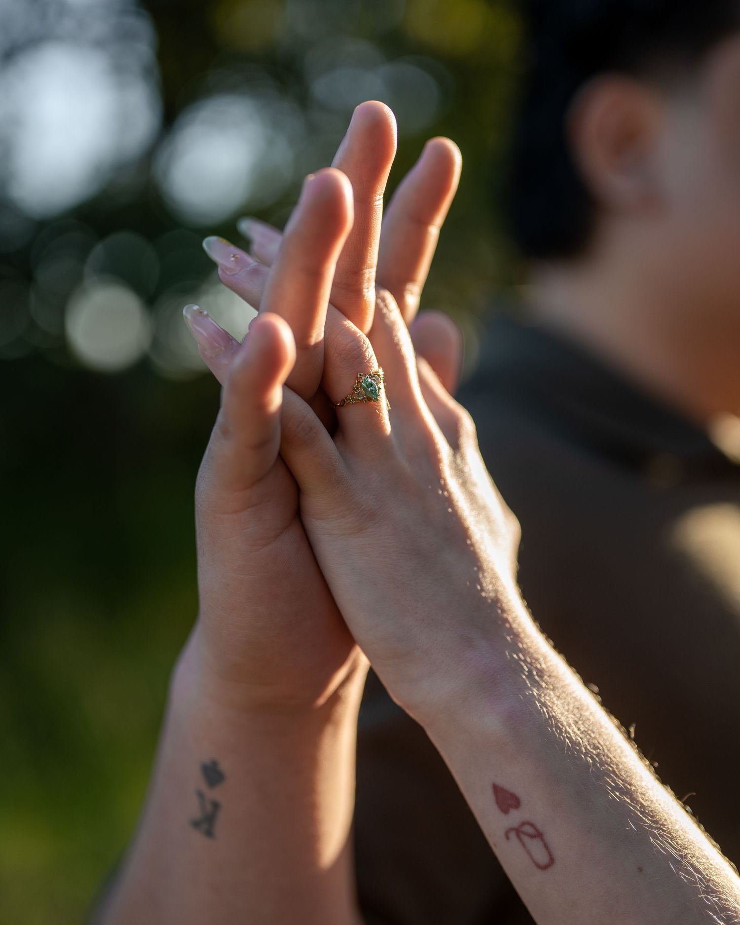 Two hands with tattoos and a green gemstone ring touch fingertips outdoors in soft golden light.