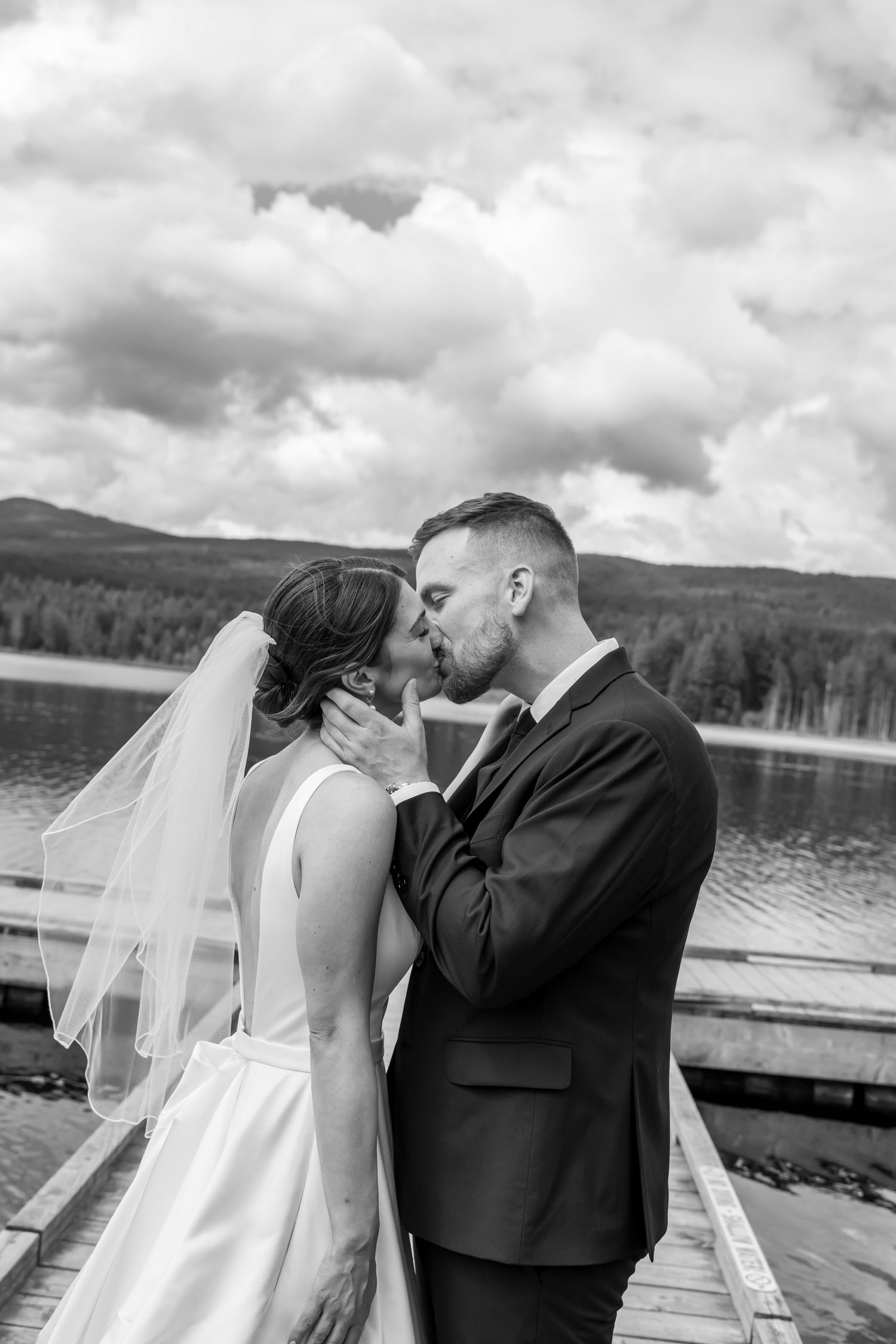 Black and white photo of a bride and groom sharing a kiss on a dock by a lake with mountains in the background.