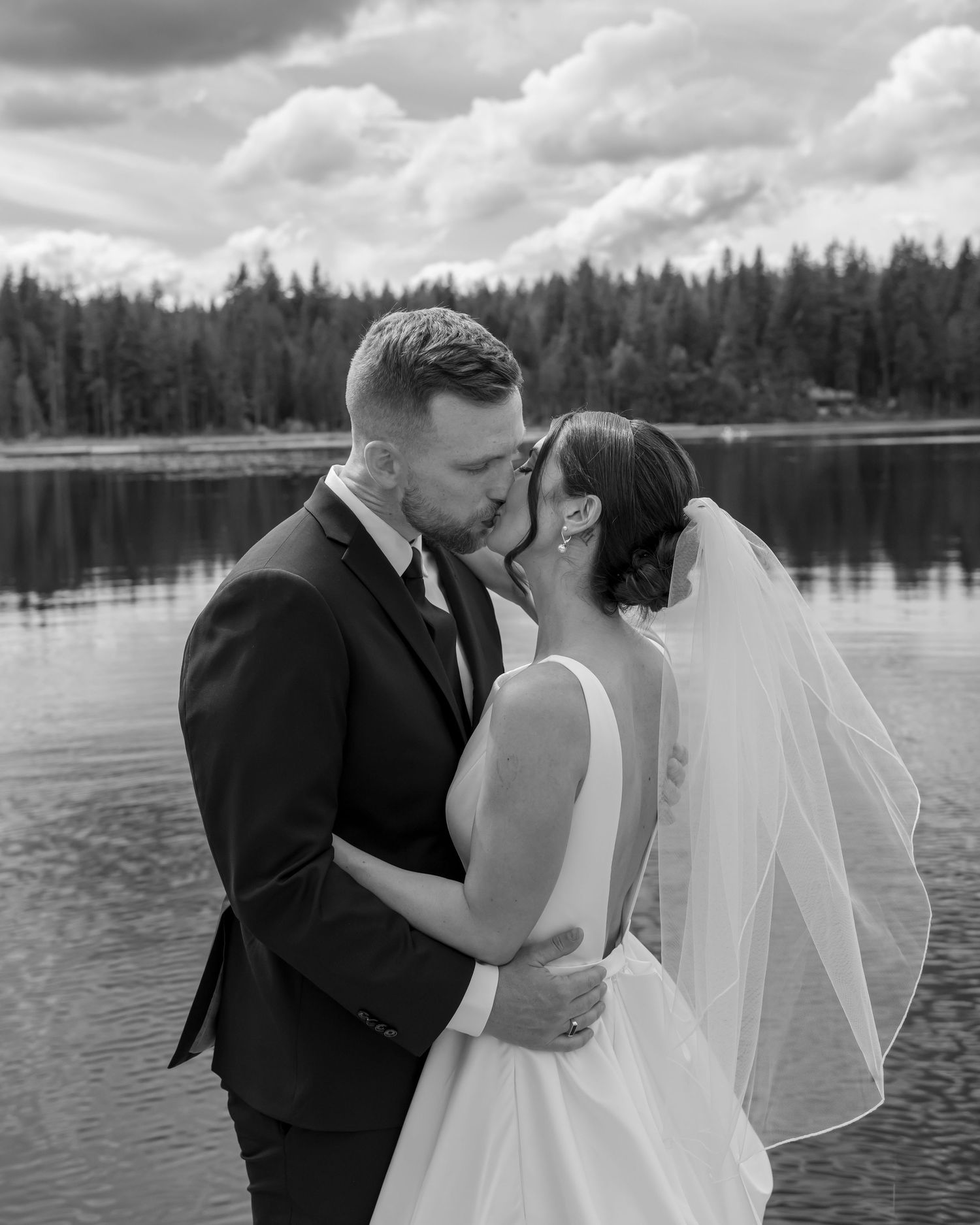 Black and white photo of a couple kissing in wedding attire by a lake surrounded by trees.