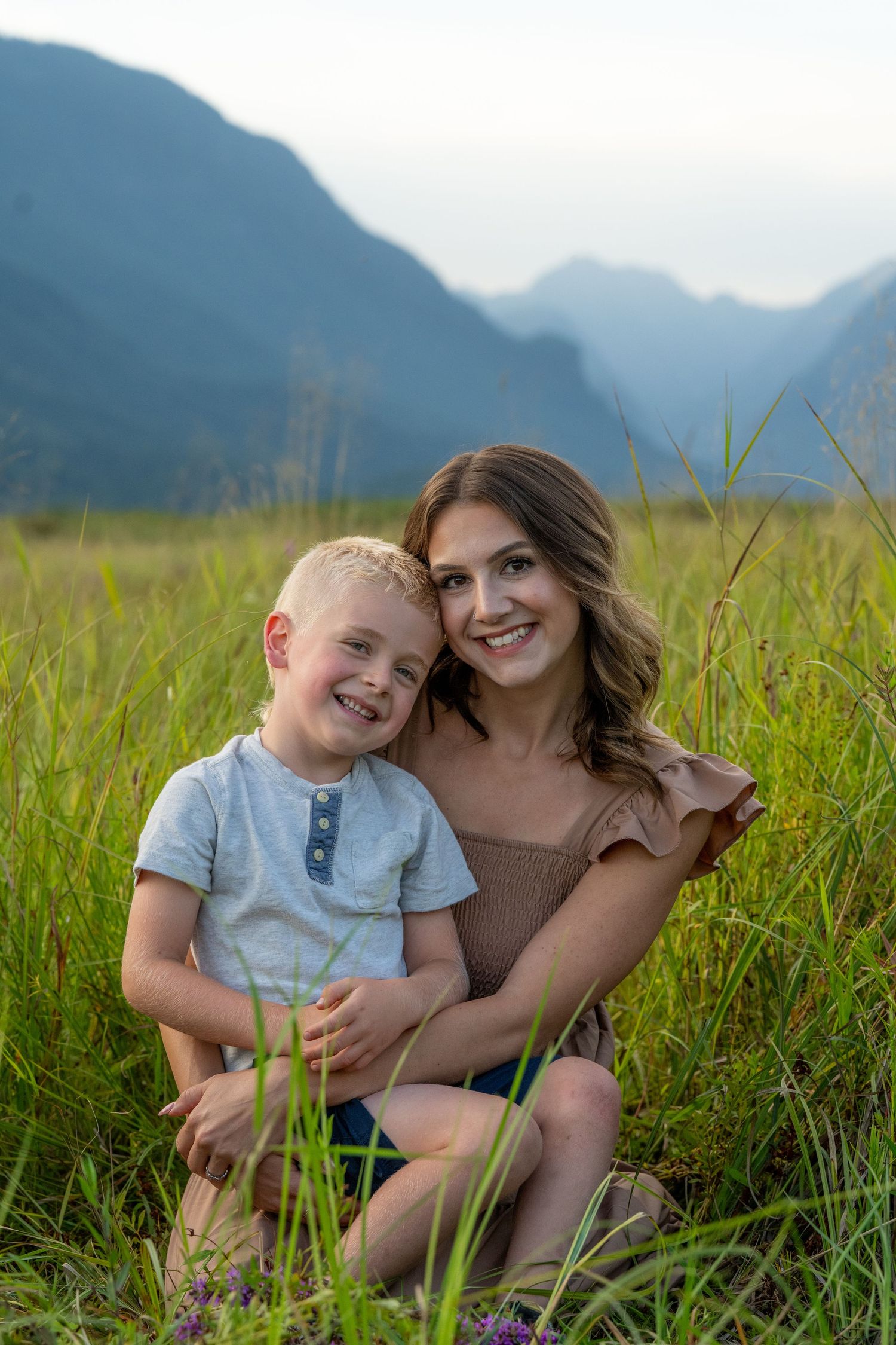 A young woman and a smiling blond boy sit together in a lush green field with misty mountains in the background.