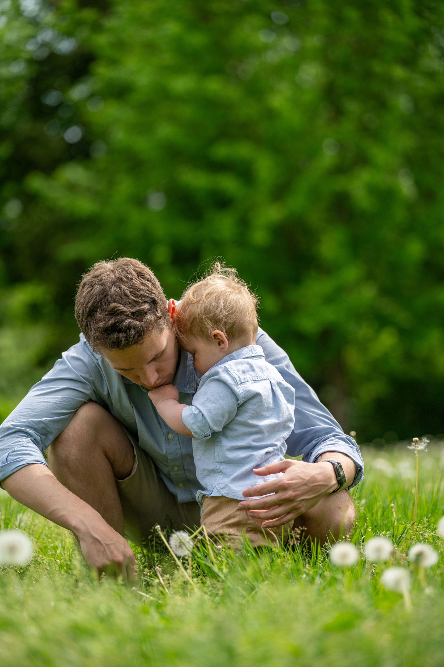 Father crouching in green meadow with dandelions, tenderly embracing his baby toddler on a sunny day.