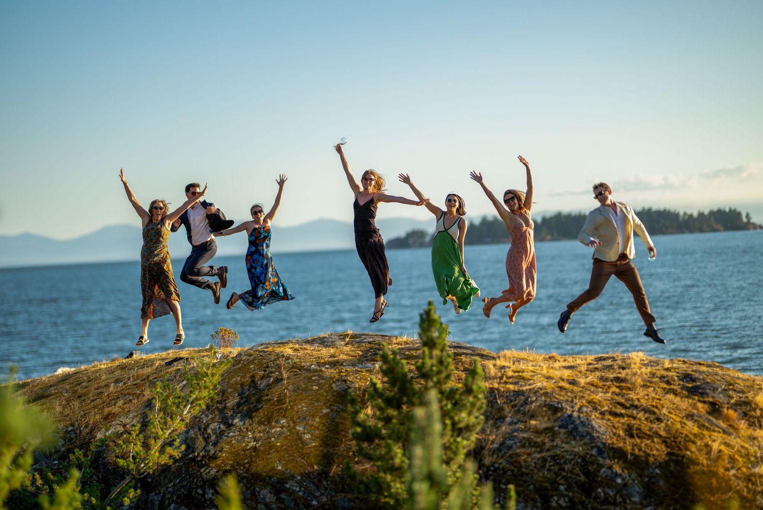 Wedding party jumps joyfully on rocky coastal outcrop with ocean backdrop.