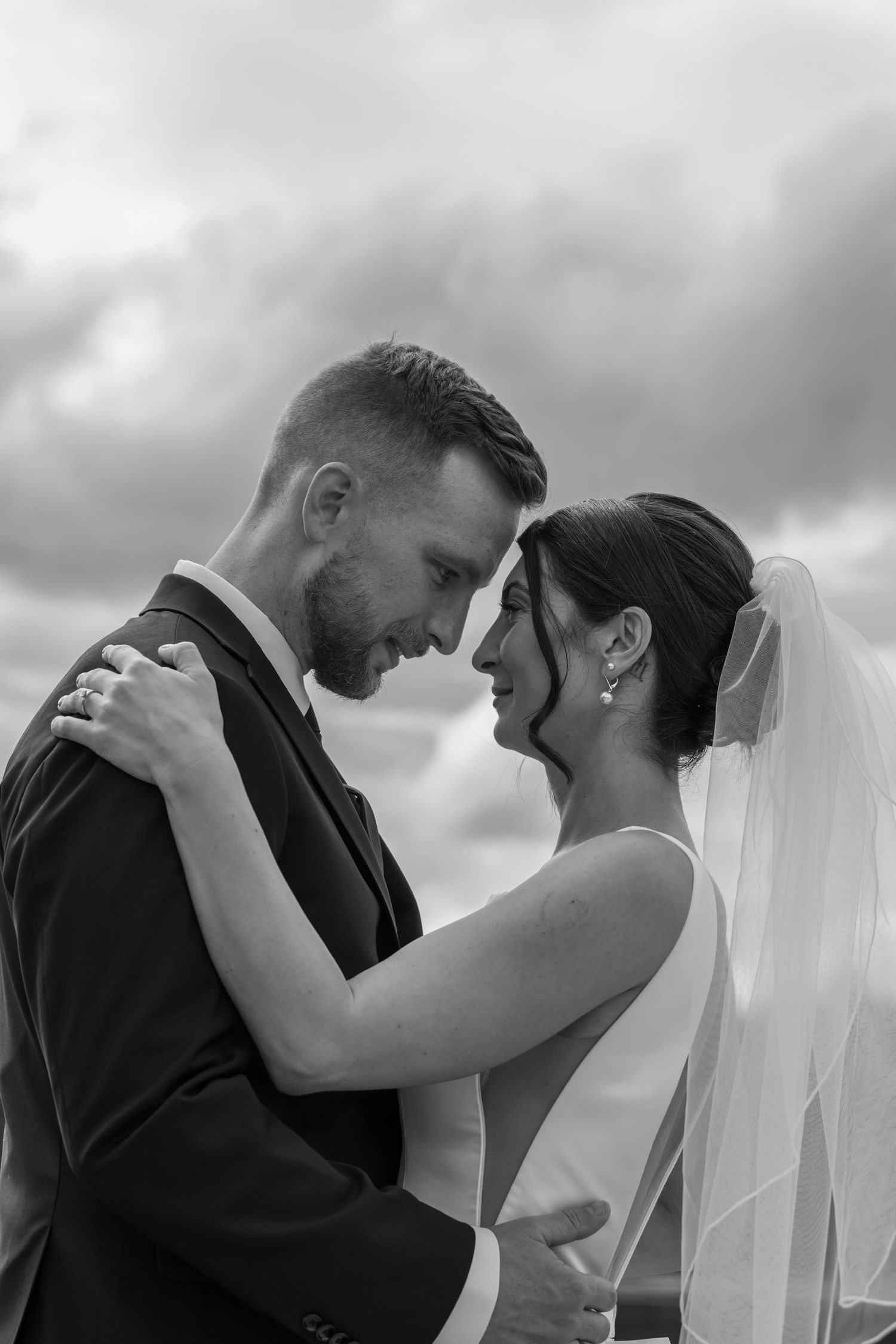 Black and white photo of a smiling bride and groom embracing outdoors under a cloudy sky on their wedding day.