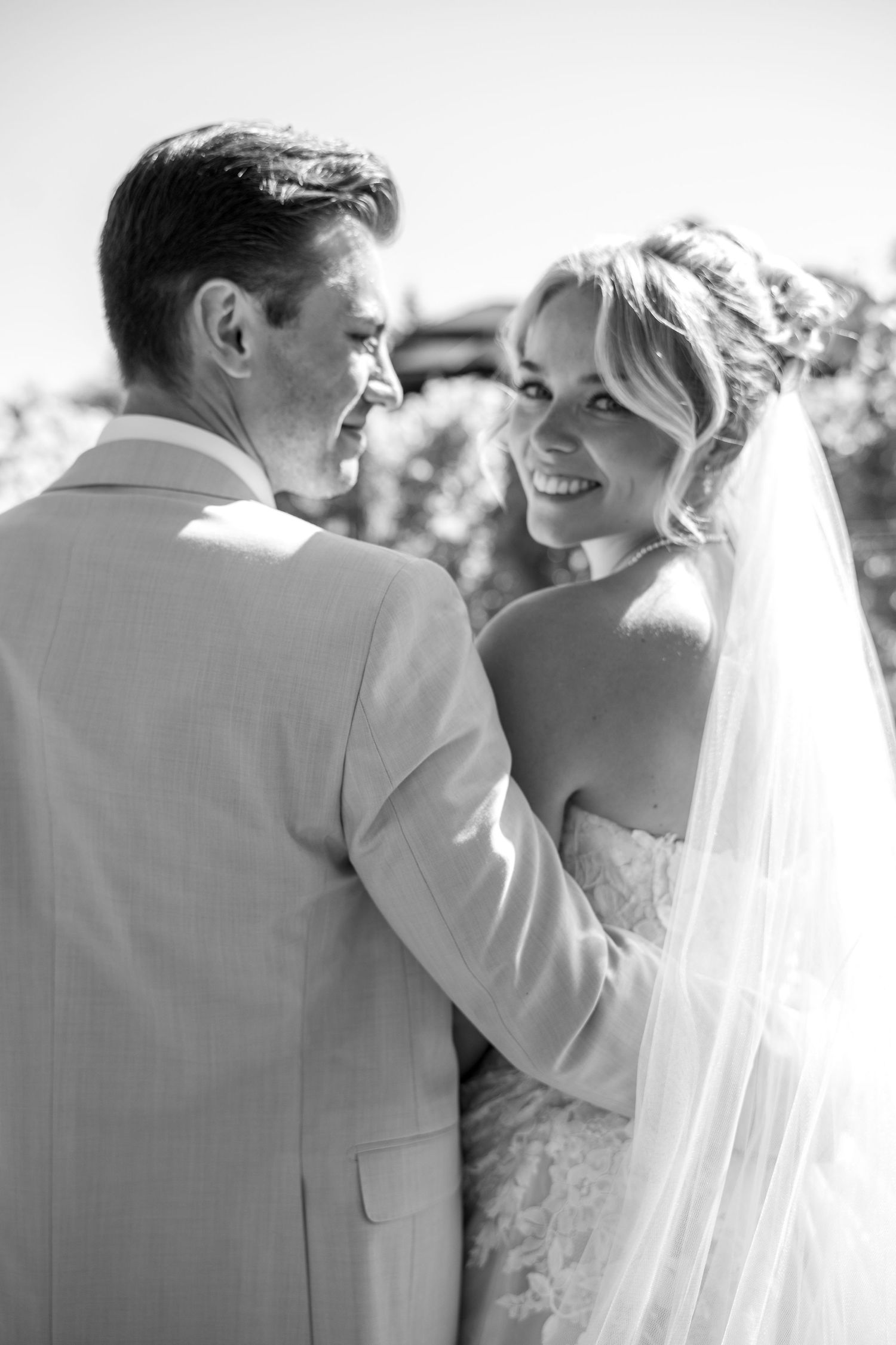 Black and white photo of a smiling bride in veil embracing her groom in a light suit at their wedding.