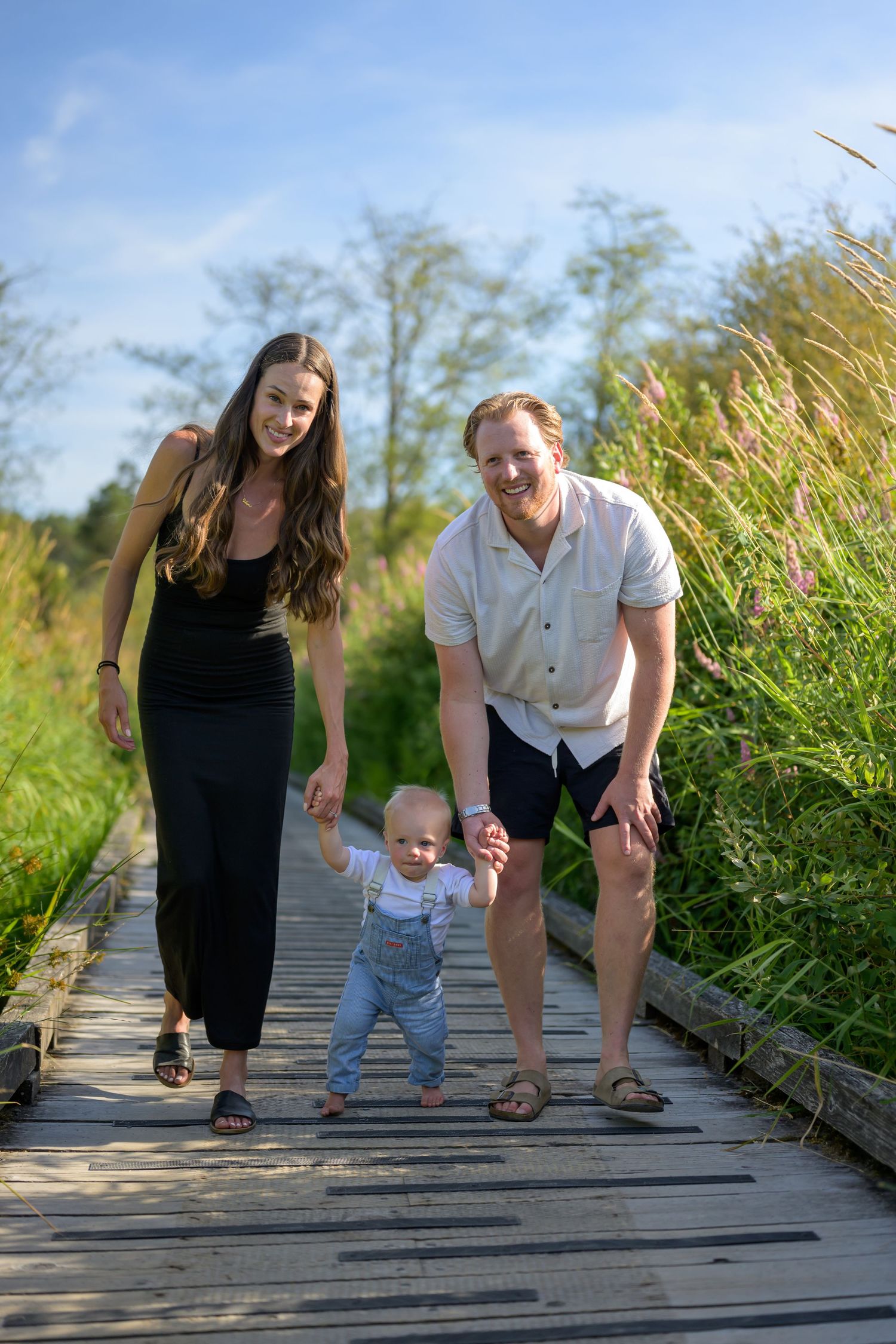 Family helping toddler take first steps on wooden boardwalk surrounded by tall grass.