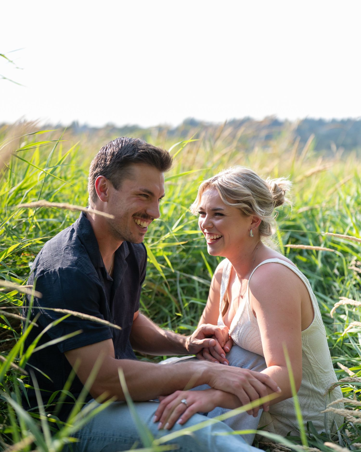 A couple lies together in tall grass during a sunny outdoor photoshoot.