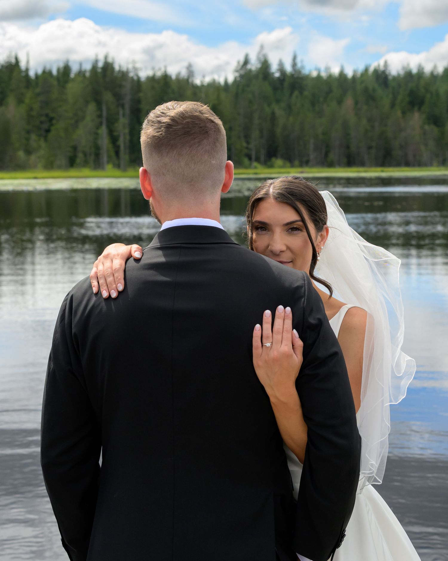 Bride smiles over groom's shoulder showing wedding ring by a serene lake surrounded by pine trees.