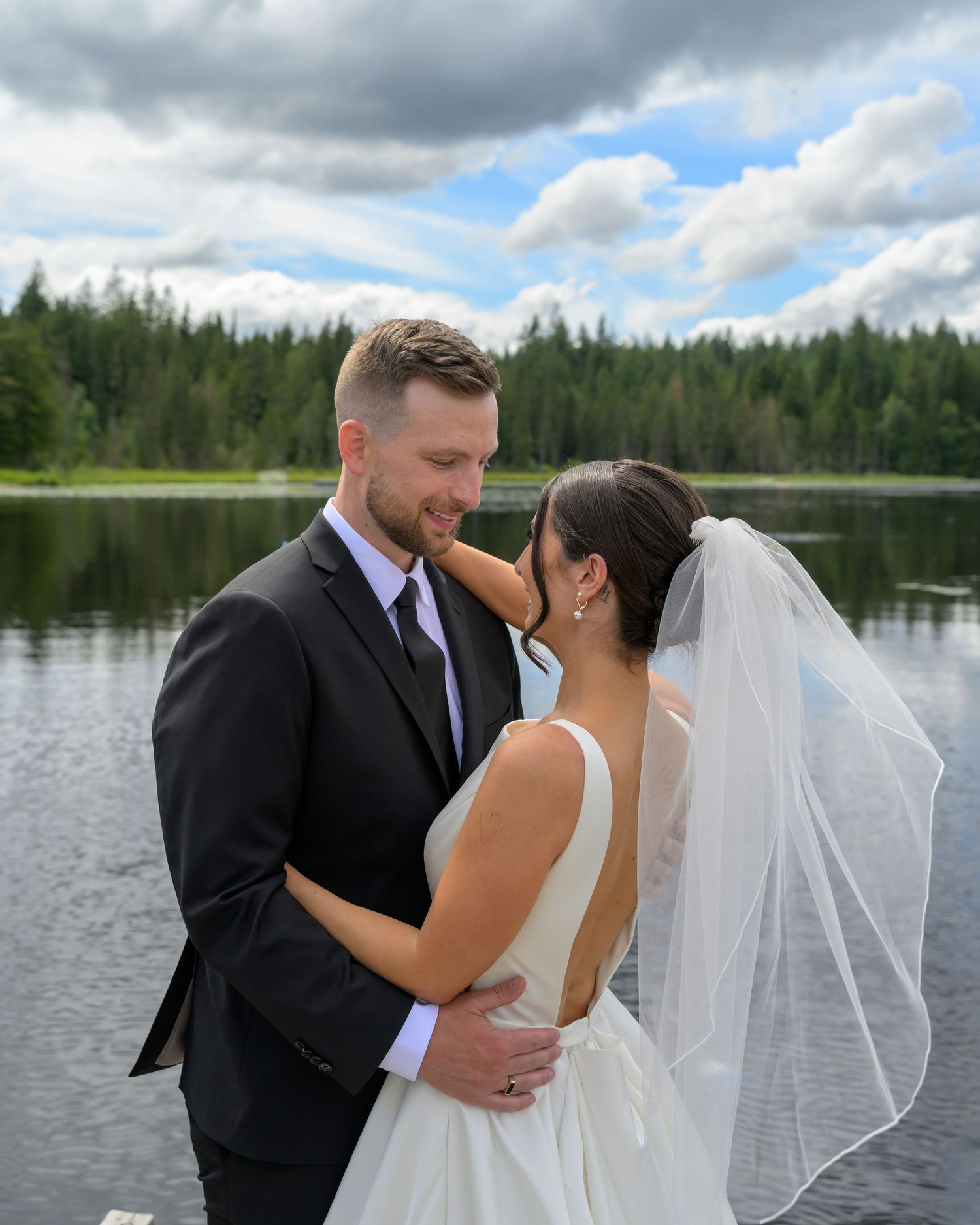 Bride and groom embrace near a lakeside forest backdrop on their wedding day.