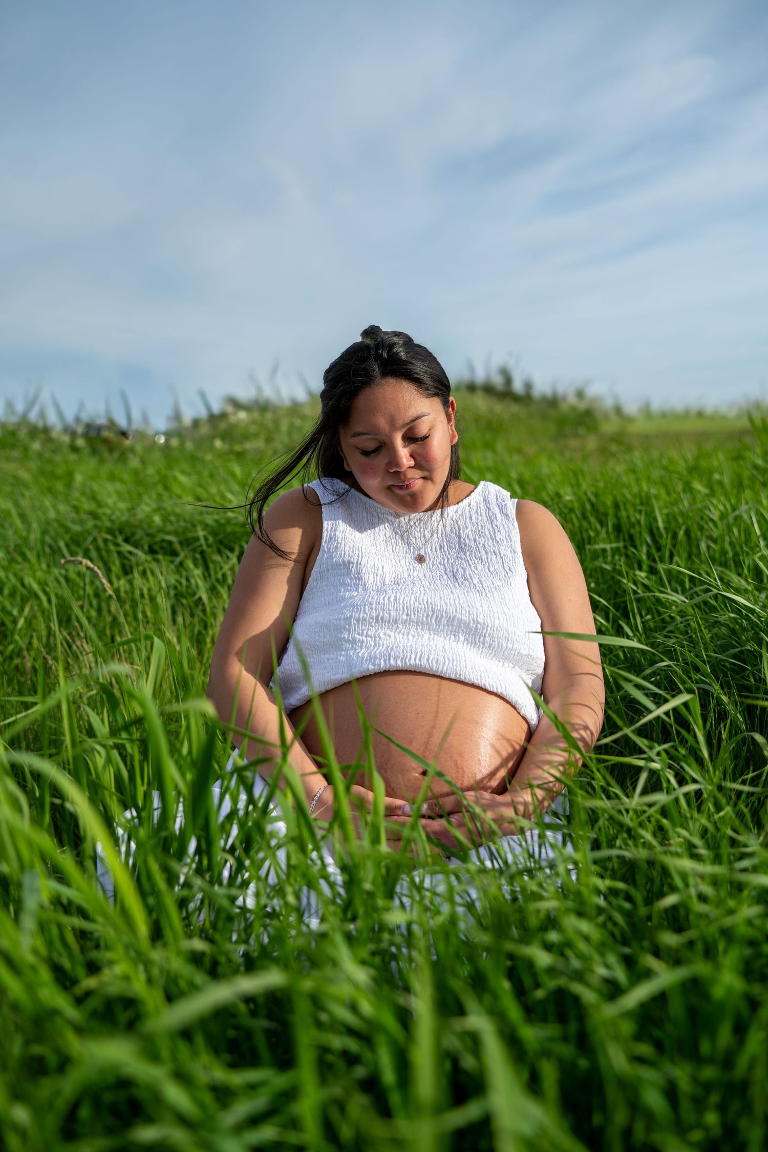 Pregnant woman in white top sitting peacefully in tall green grass, cradling her baby bump under a blue sky.