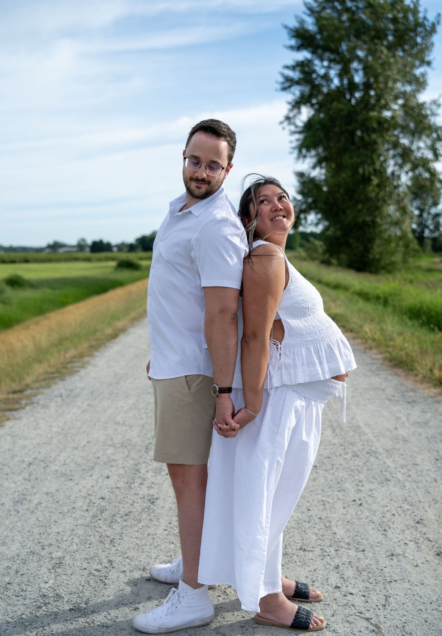 Couple in white outfits holding hands on a rural dirt road, woman visibly pregnant, surrounded by green fields.