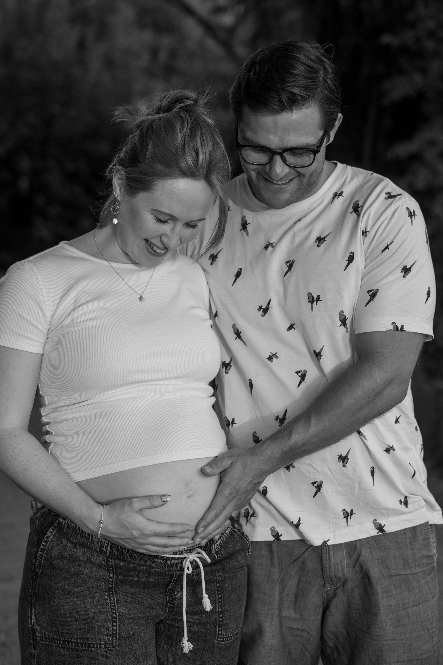 Black and white photo of a smiling pregnant couple tenderly cradling the baby bump outdoors.