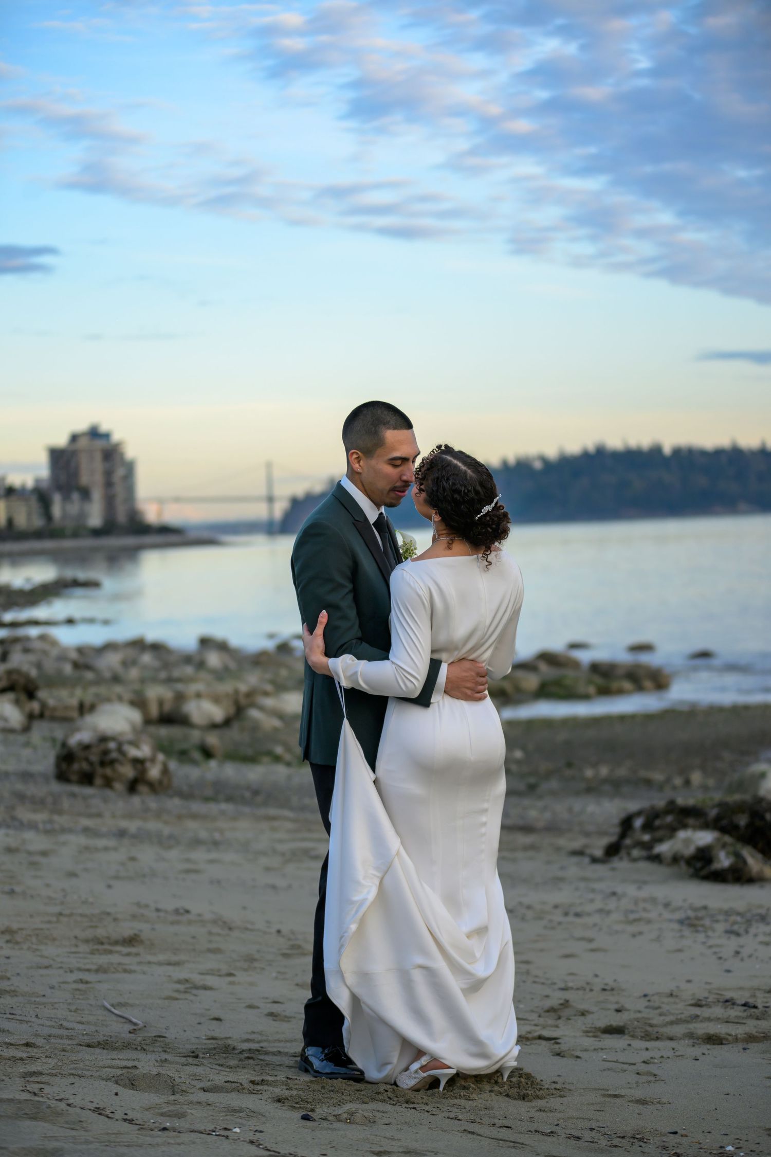 A newlywed couple embraces on a beach with water and buildings in the distance during golden hour.