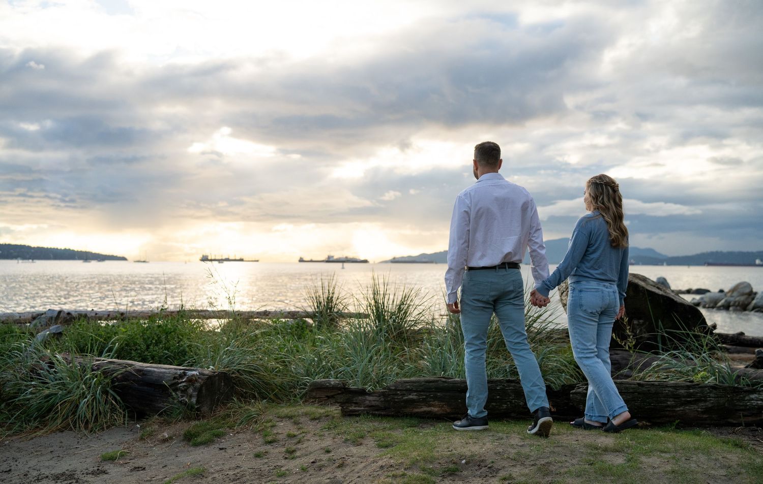 Couple holding hands watching sunset over calm ocean waters with dramatic cloudy sky from rocky shoreline.