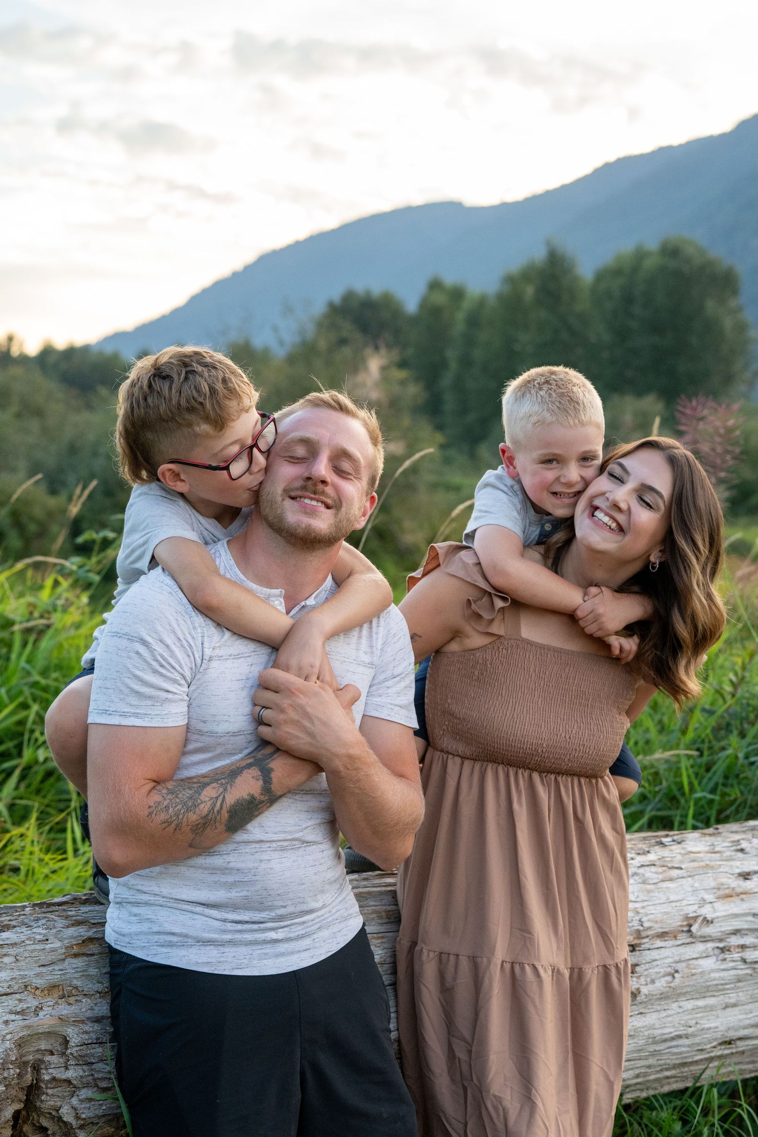 A happy family of four poses outdoors, two young boys hugging their smiling parents against a scenic mountain backdrop.