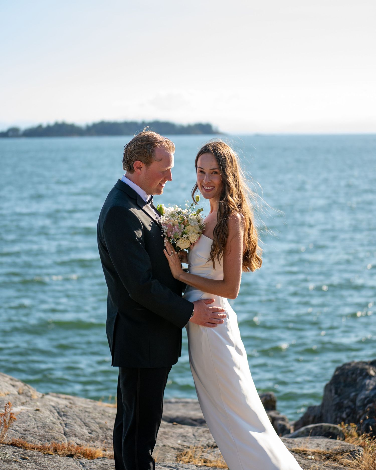 Romantic coastal wedding photo shoot with couple embracing on rocky shoreline by the sea.