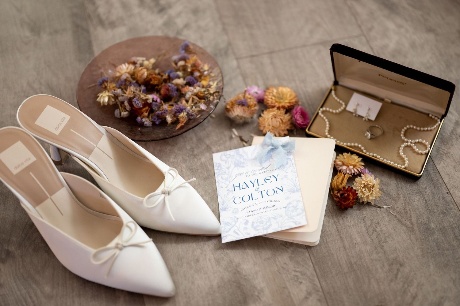 White bridal heels, wedding invitation for Hayley and Colton, pearl jewelry, and dried floral accents arranged on a wooden floor.