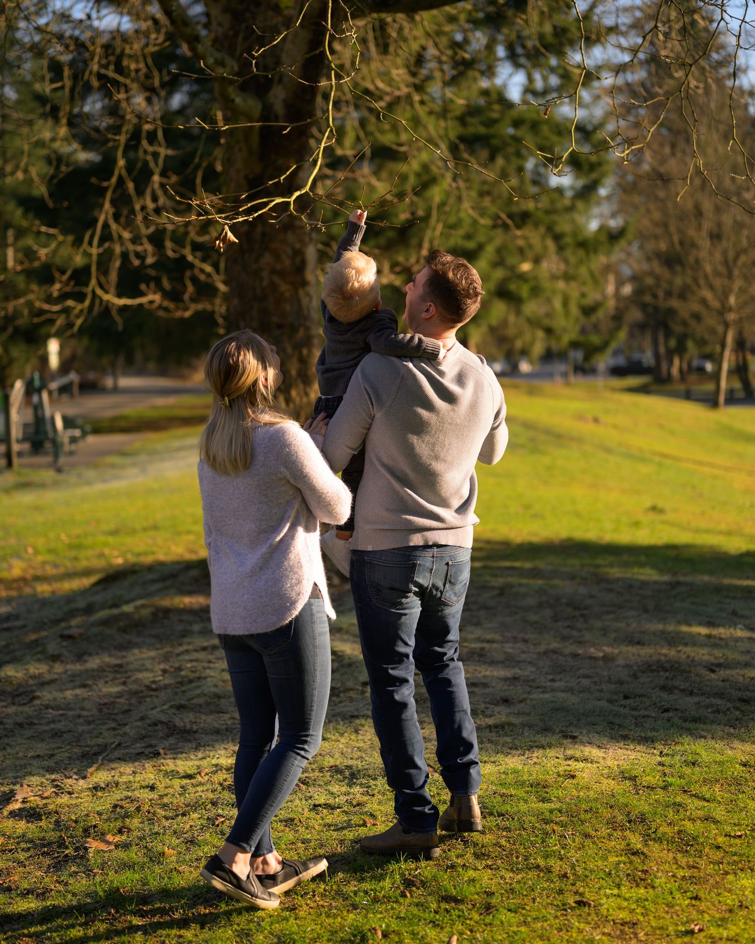 Parents and their toddler walk together on a grassy hill during a sunny morning in Gates Park, Port Coquitlam