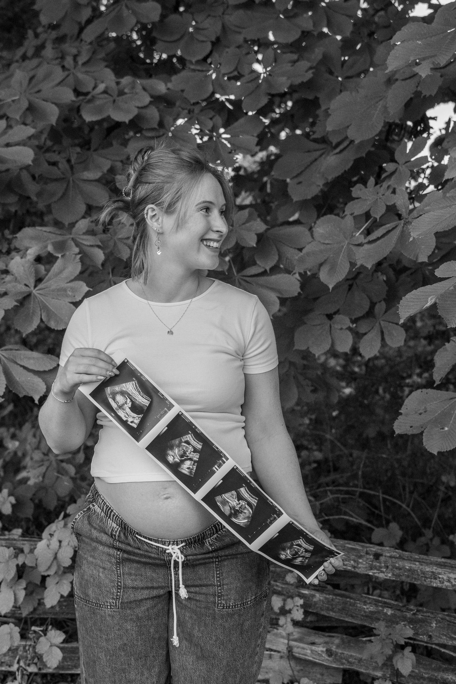 Black and white photo of smiling pregnant woman holding ultrasound strip outdoors near lush foliage.