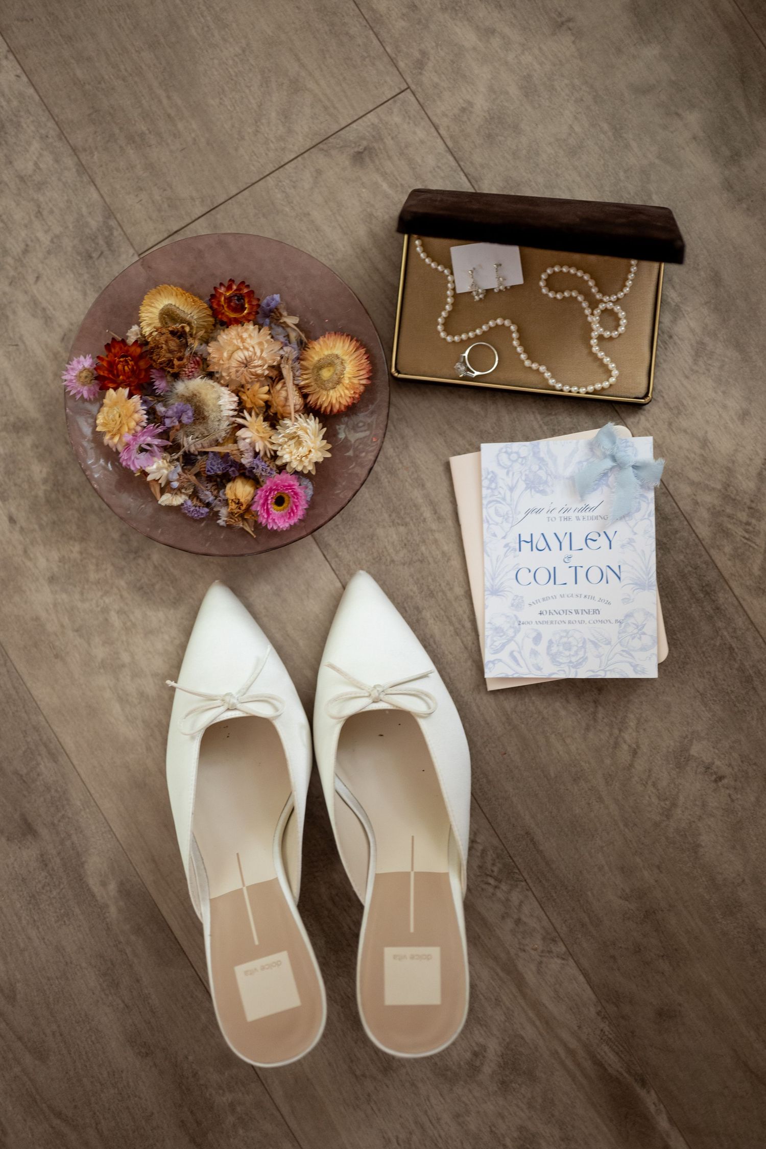Flat lay of white bridal shoes, dried flowers, pearl jewelry box, and Hayley & Colton wedding invitation on wood floor.