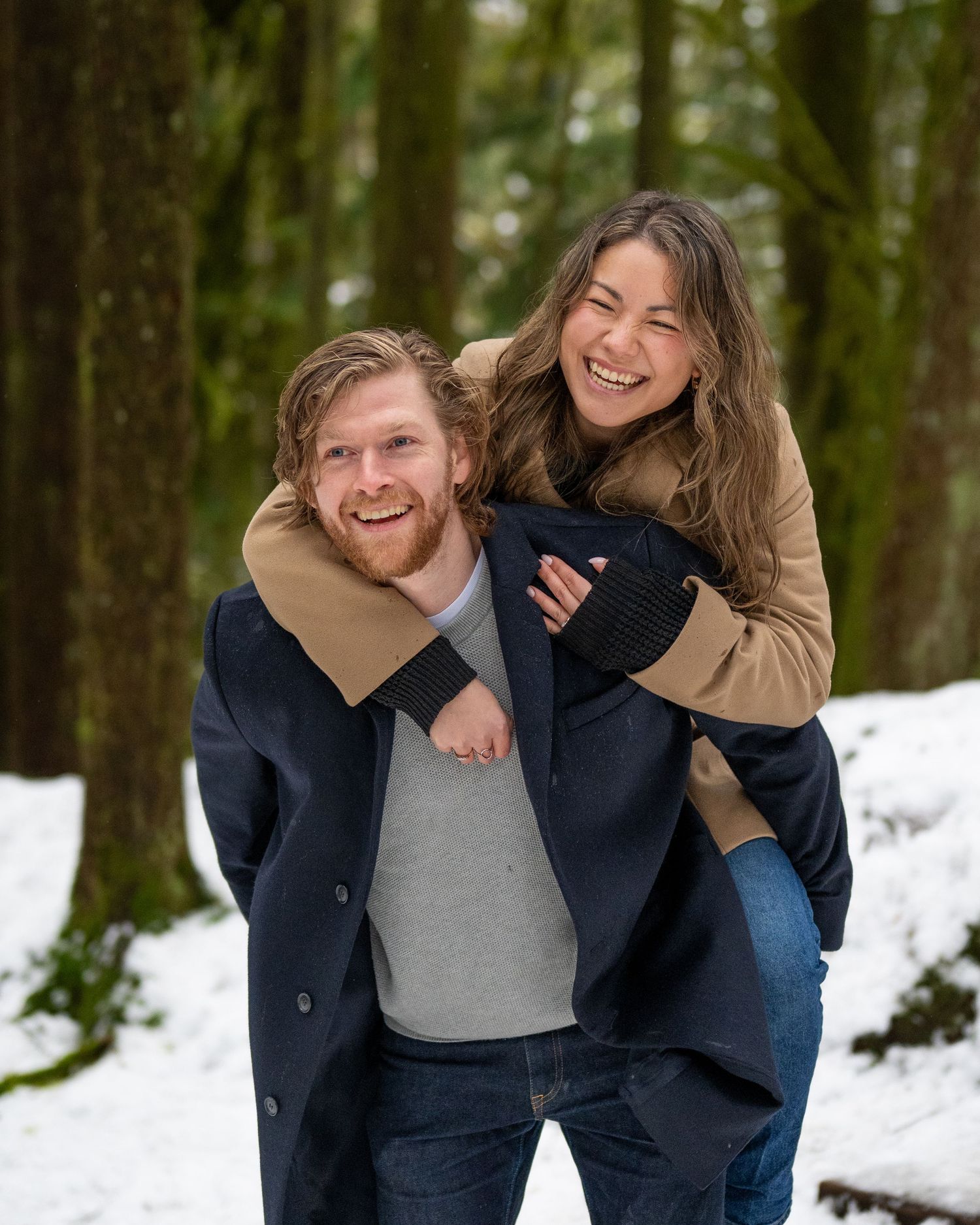 Playful couple laughing together in snowy forest wearing winter coats.