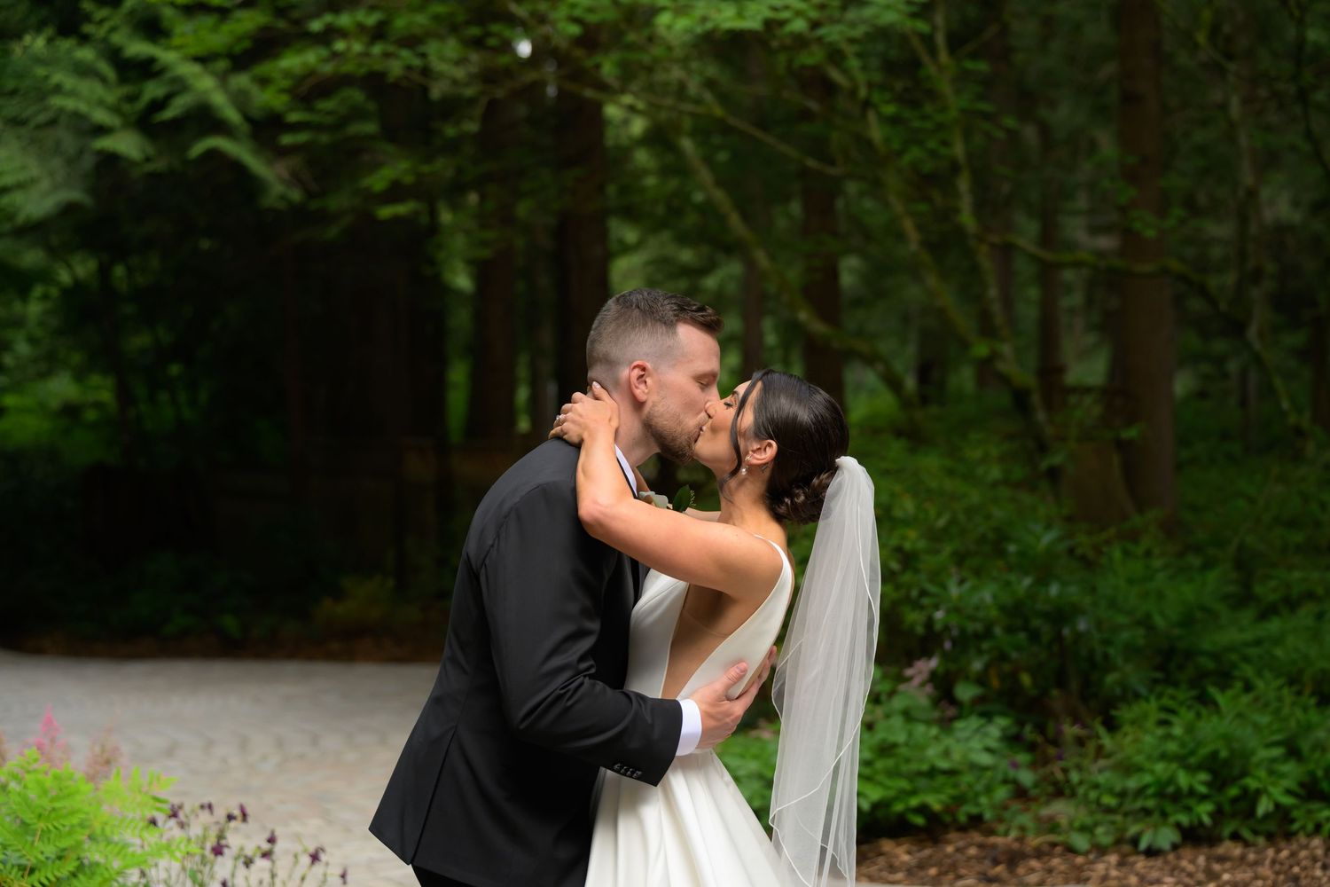 Bride and groom share a romantic kiss on a garden path surrounded by lush green trees on their wedding day.