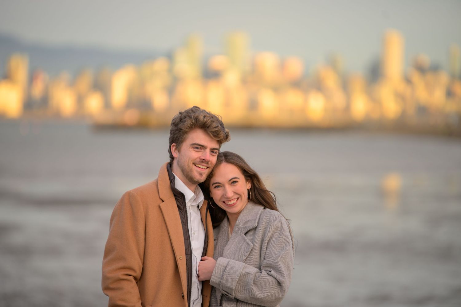 Couple sharing romantic moments at sunset by the water with city skyline in background.