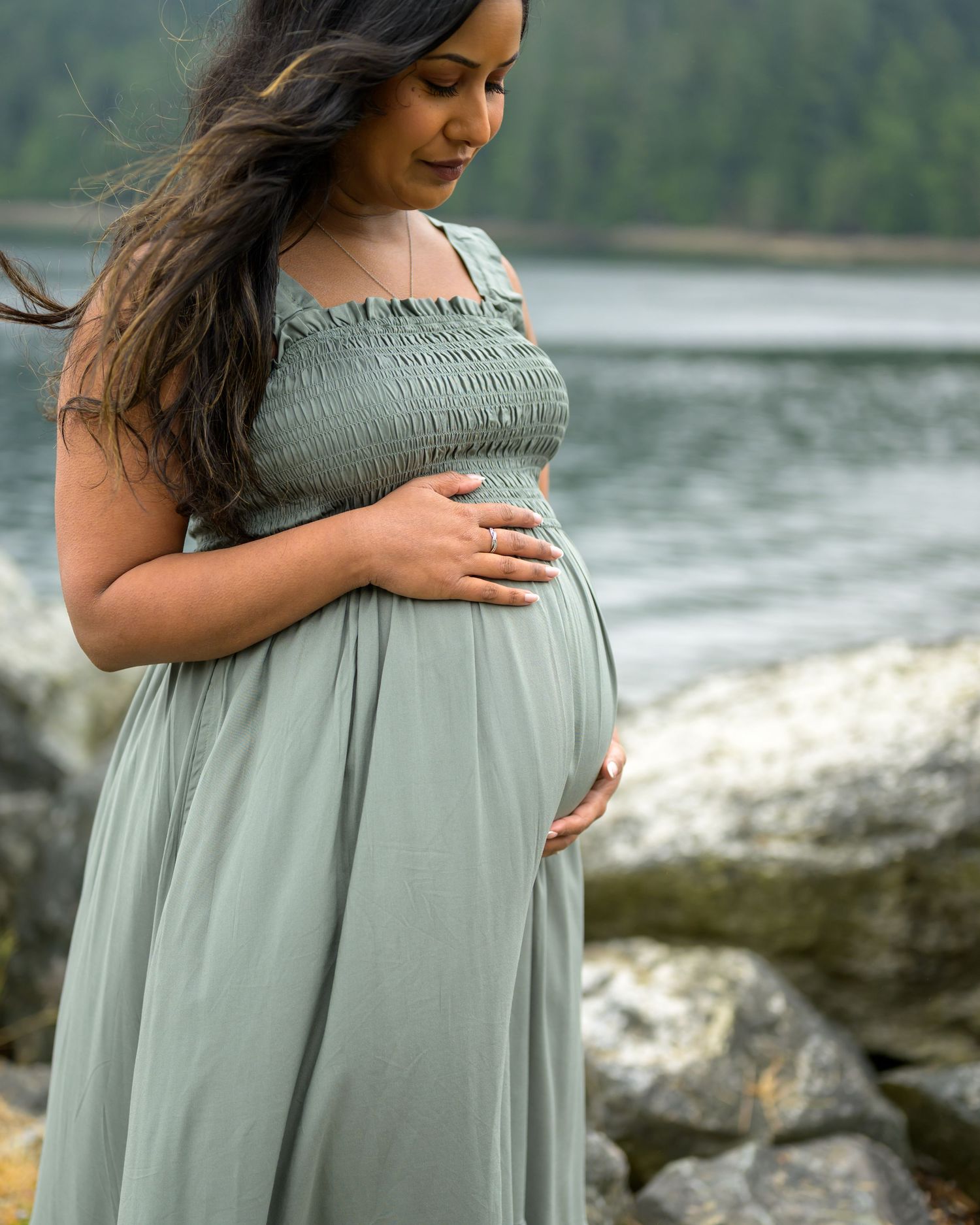 Pregnant woman in sage green smocked maxi dress cradling baby bump near rocky shoreline.