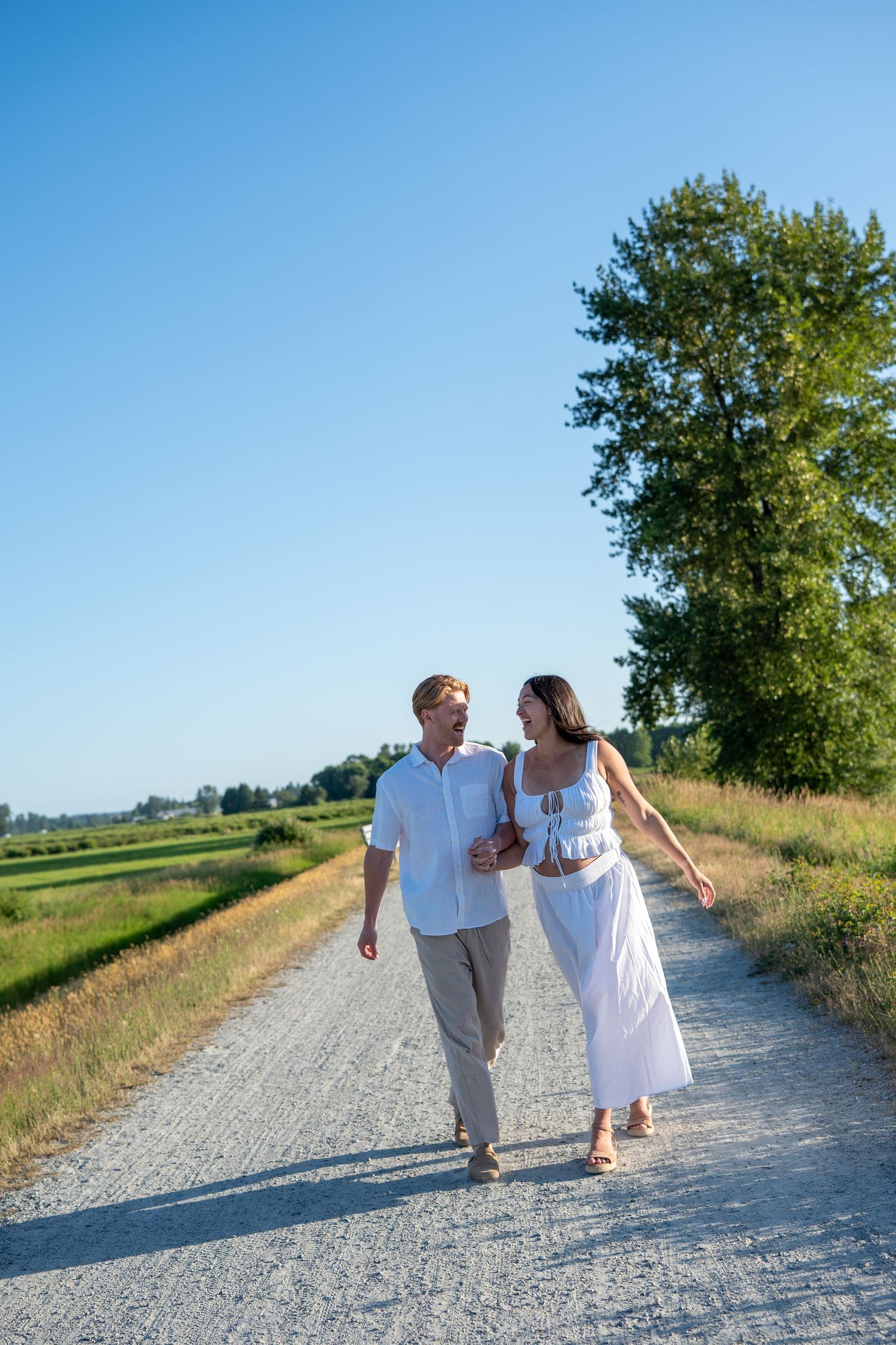 Couple in white outfits walking together on a sunny rural gravel path surrounded by green trees and fields.