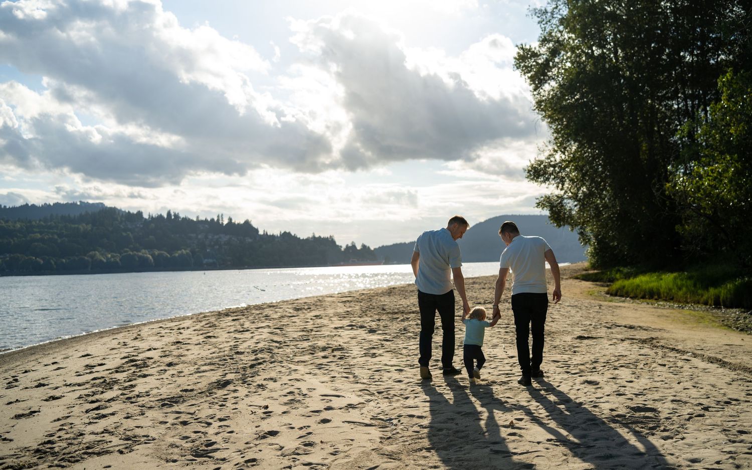 Two parents walk with a young child on a sandy beach beside a calm river on a cloudy day.