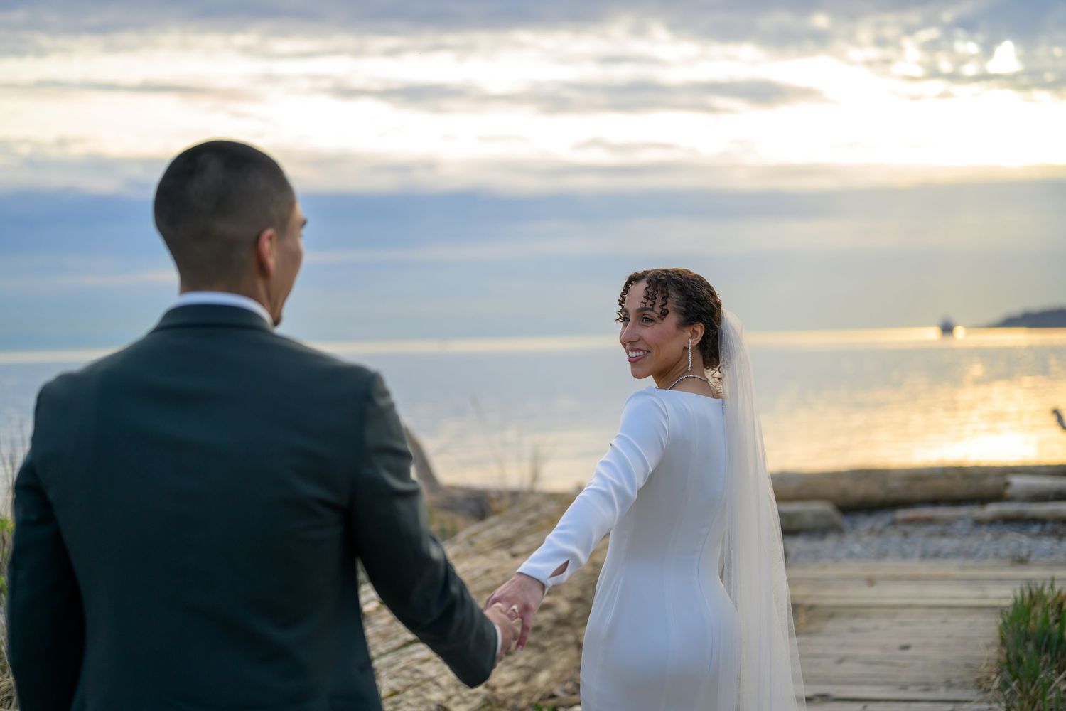 Wedding couple walking along scenic beach at sunset with cloudy sky.