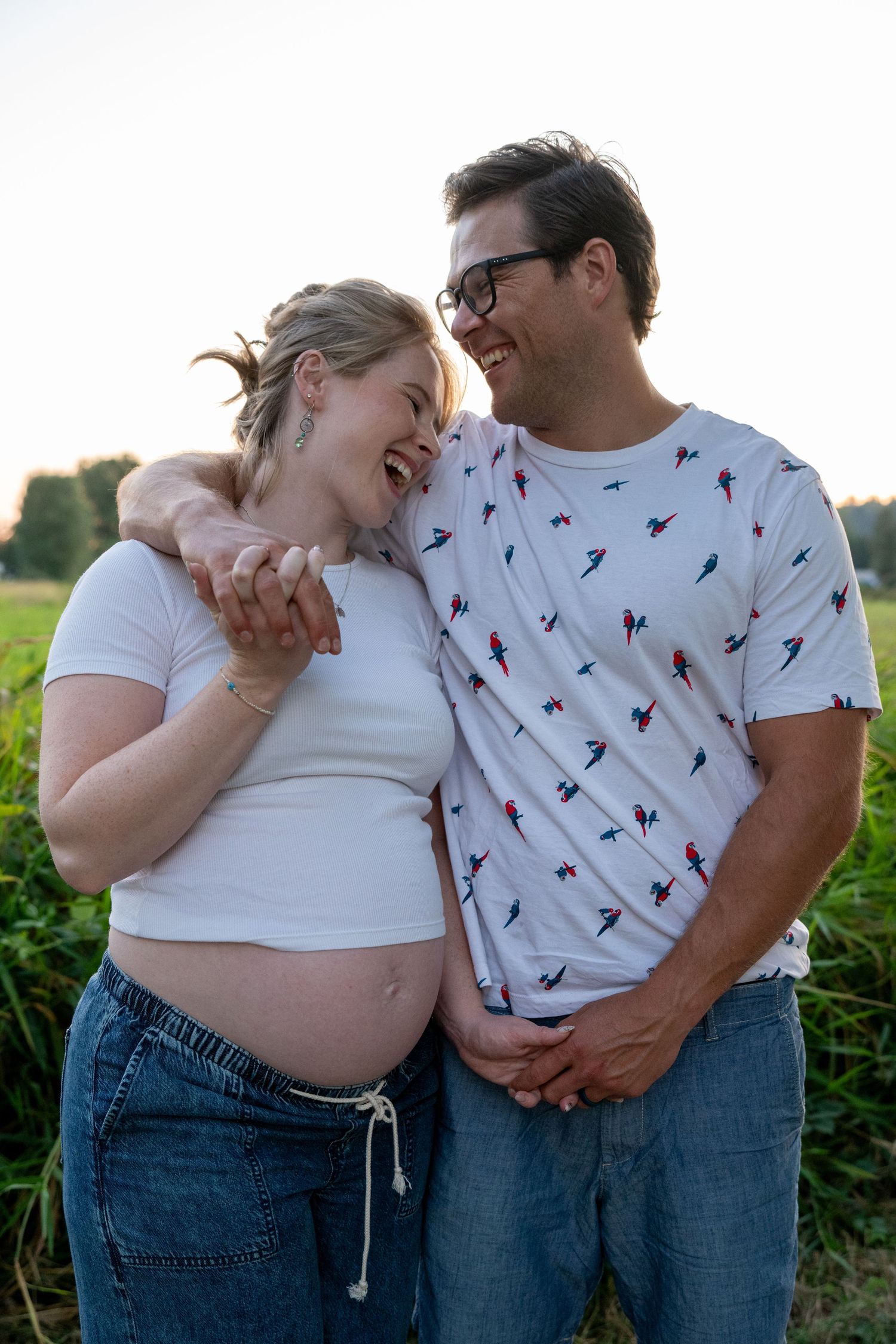 Smiling pregnant couple embracing outdoors in a green field at sunset, woman in white crop top showing baby bump.