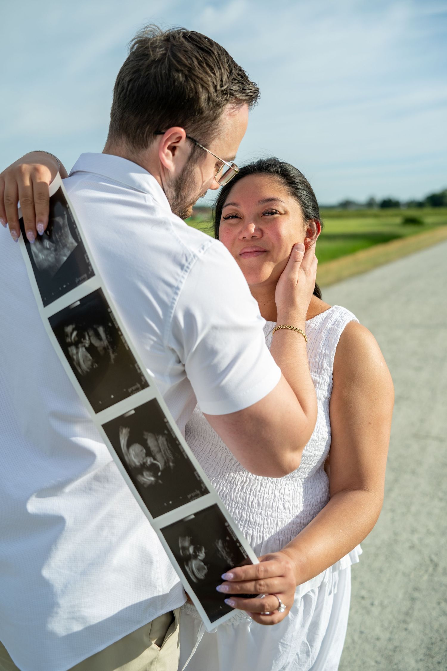 Couple embracing outdoors holding ultrasound photos, pregnancy announcement on sunny day with scenic road background.