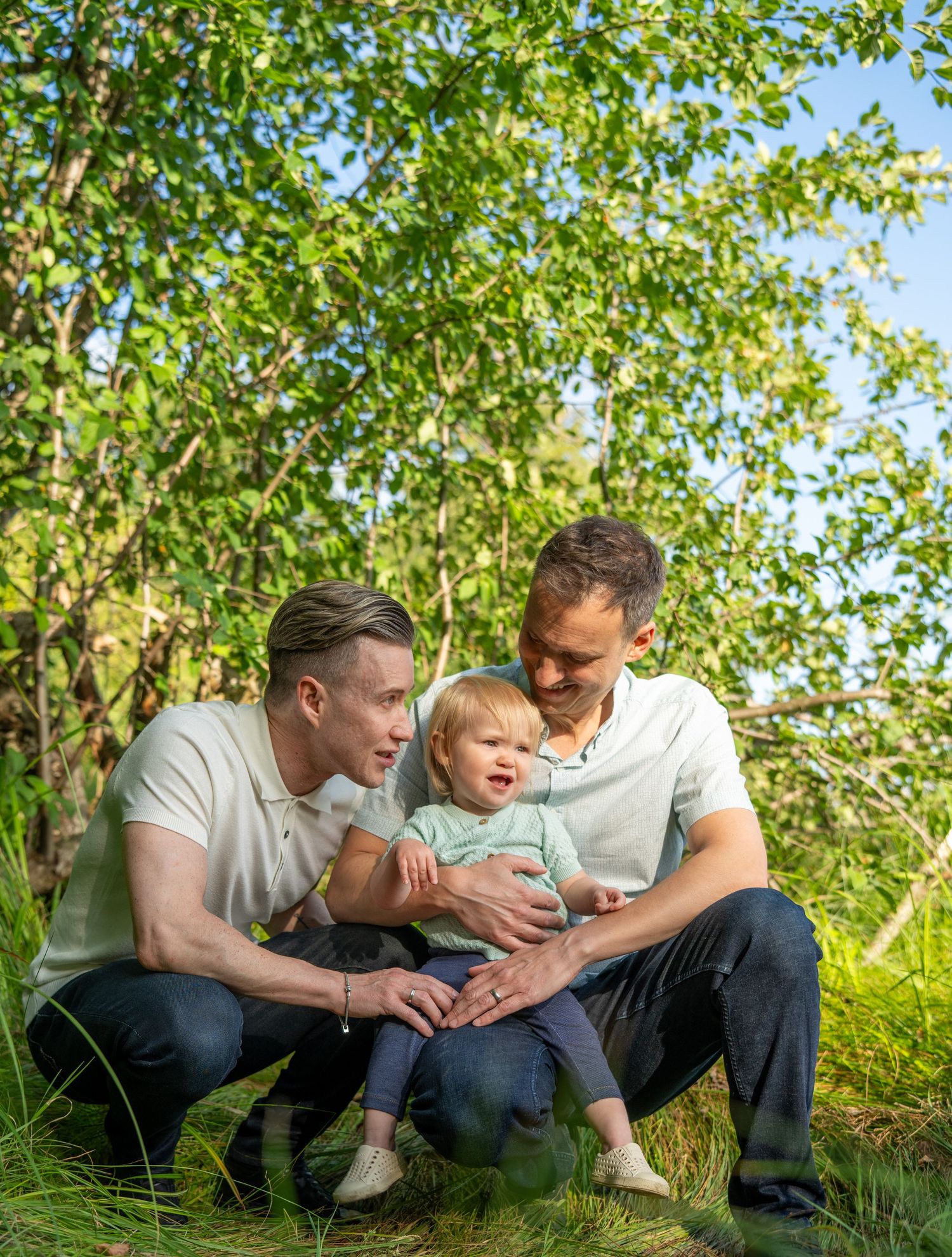 Two men crouching outdoors with a toddler between them, surrounded by lush green trees on a sunny day.