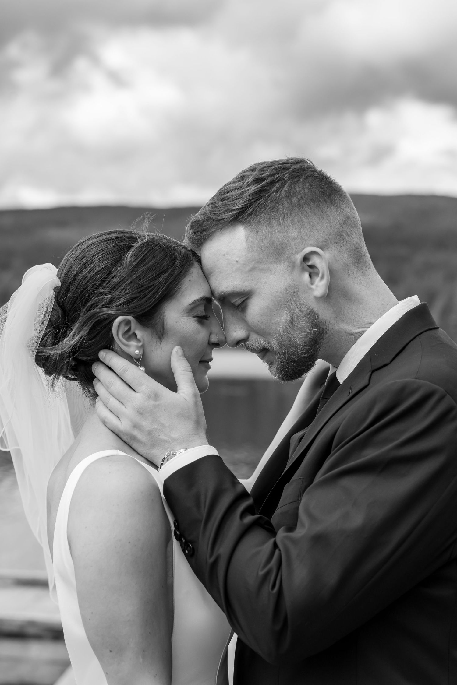 Black and white photo of a bride and groom sharing an intimate moment, foreheads touching, outdoors on their wedding day.