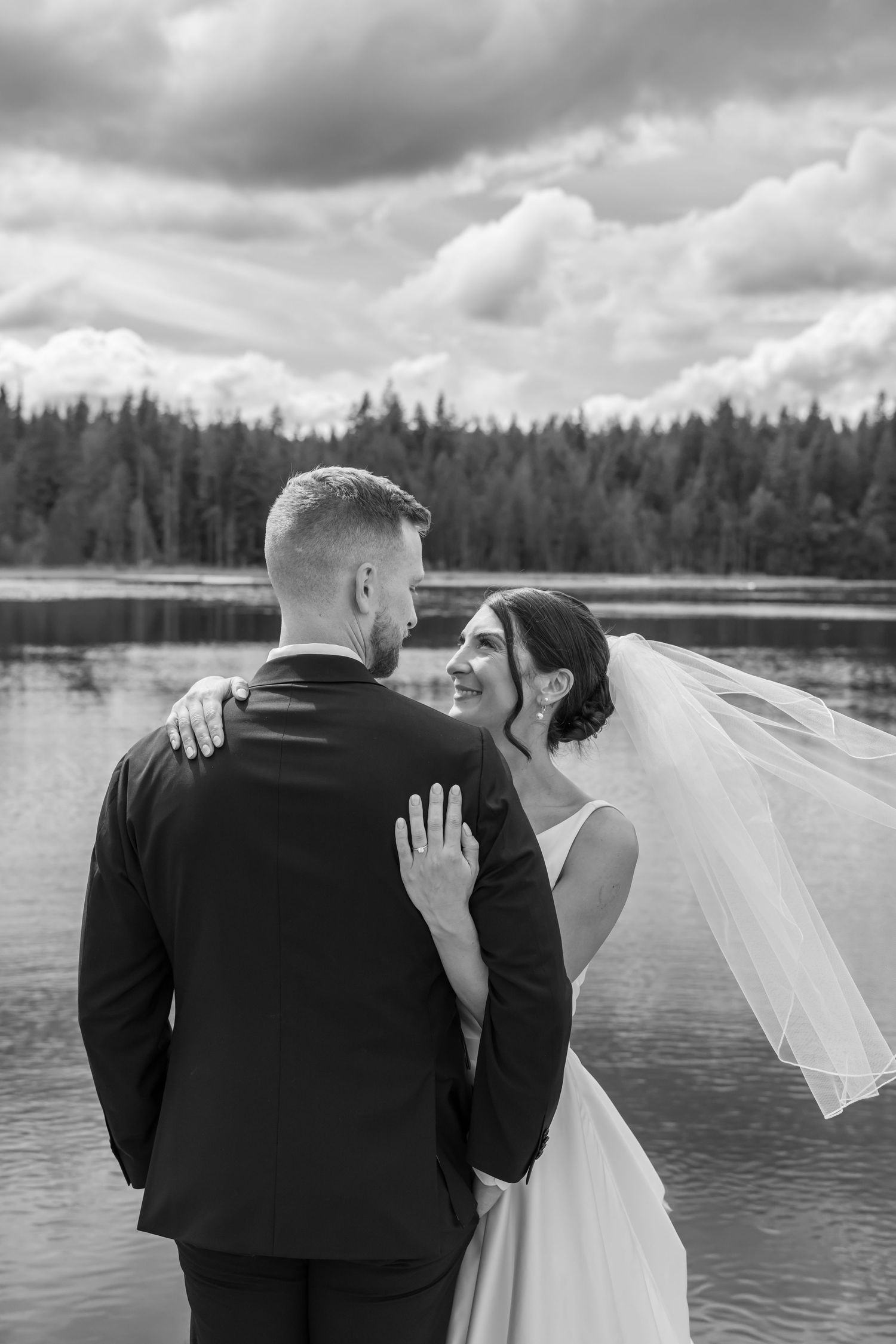 Black and white photo of bride and groom embracing by a lake with forest backdrop on their wedding day.