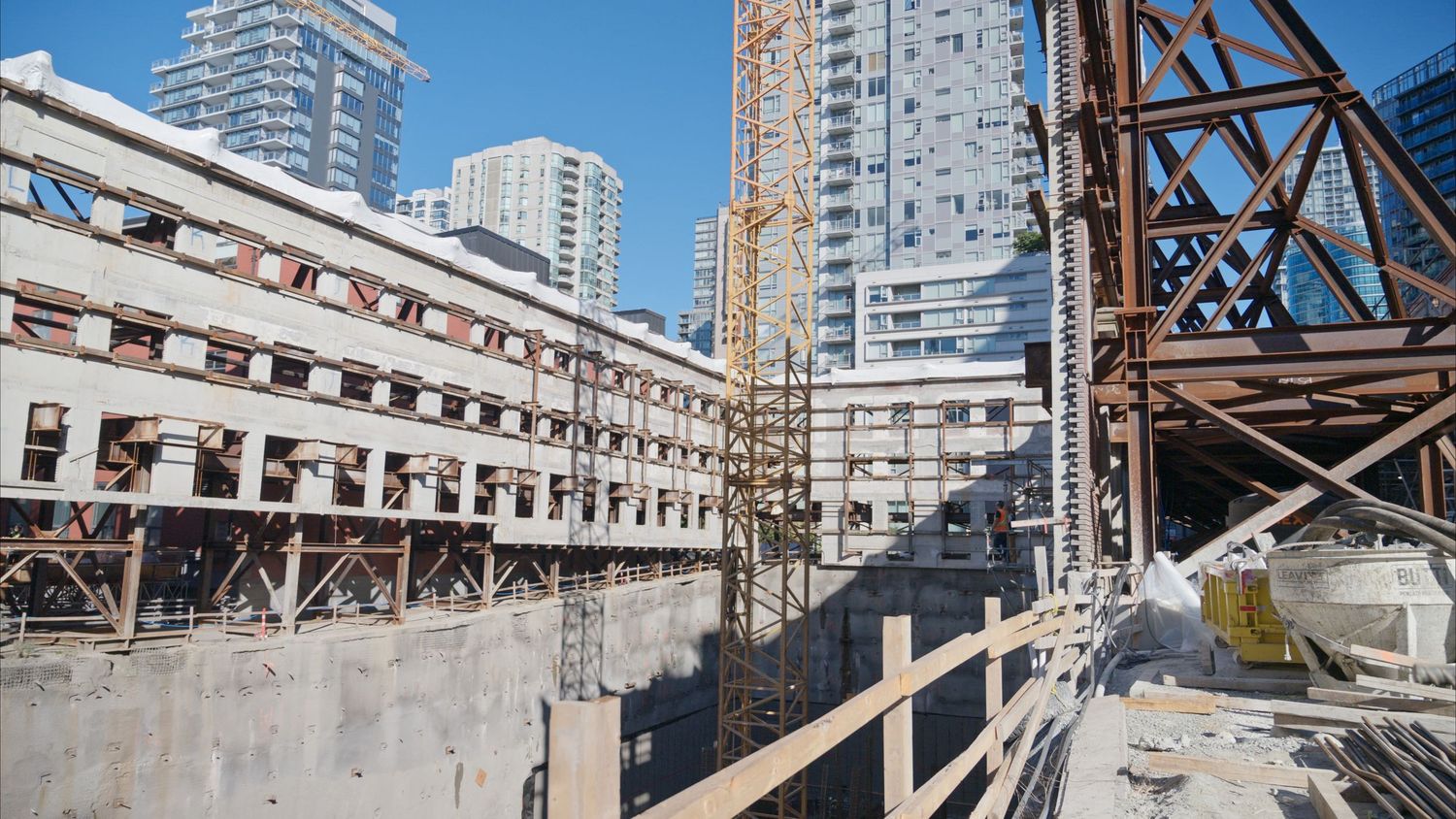 Wide panoramic view of construction site with exposed steel beams and high-rise buildings in background.