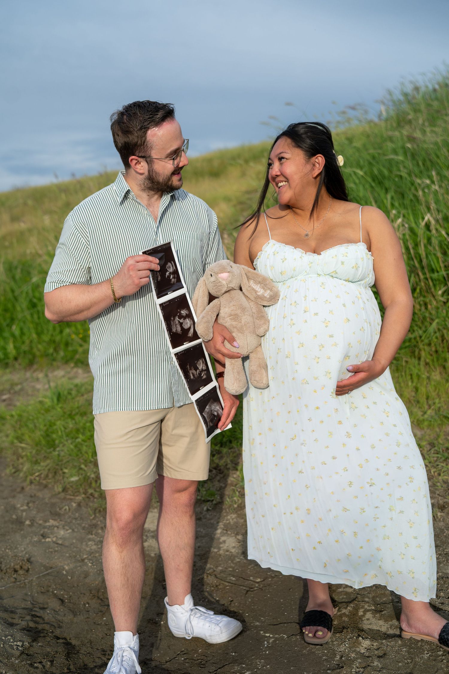 Smiling pregnant couple outdoors holding ultrasound photos and a stuffed bunny toy in a grassy natural setting.