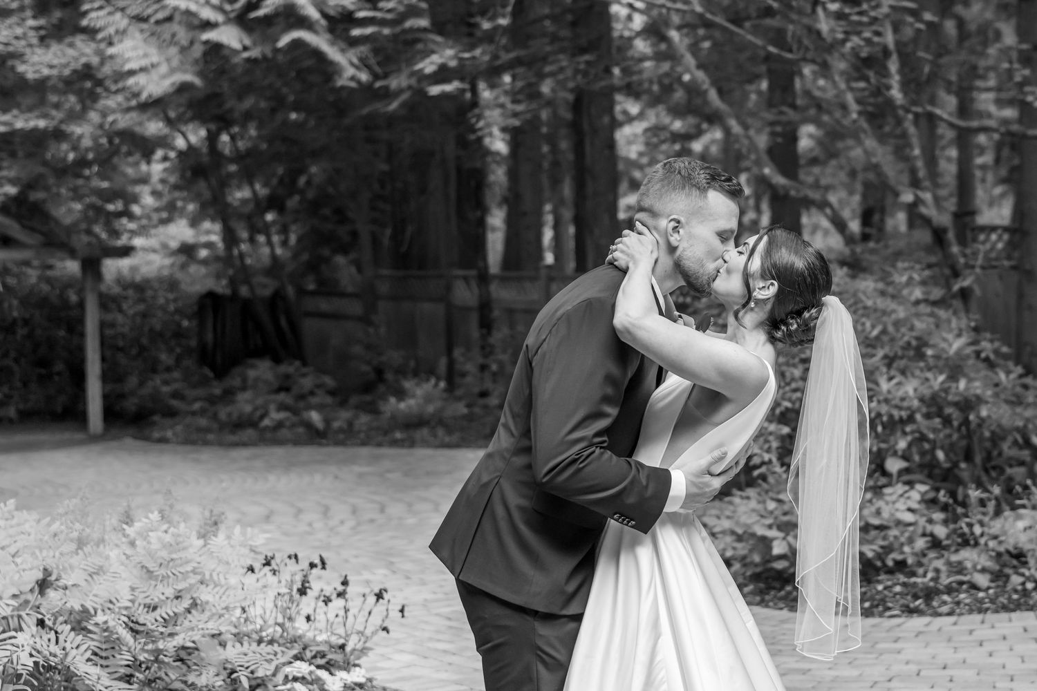 Black and white photo of a bride and groom sharing a romantic kiss outdoors surrounded by lush greenery.