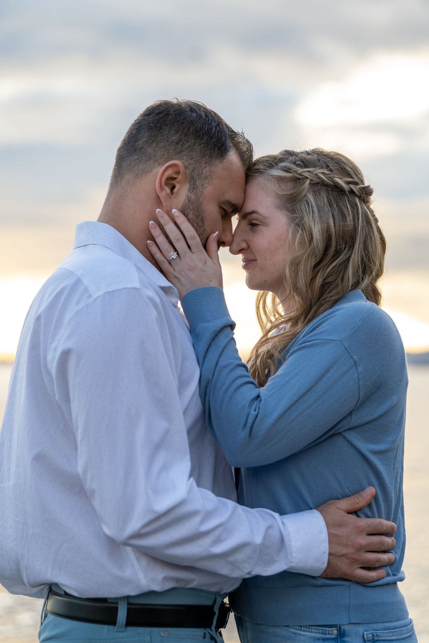 Romantic couple embracing at sunset beach, woman in blue top touching man's face, sharing an intimate forehead-to-forehead moment.