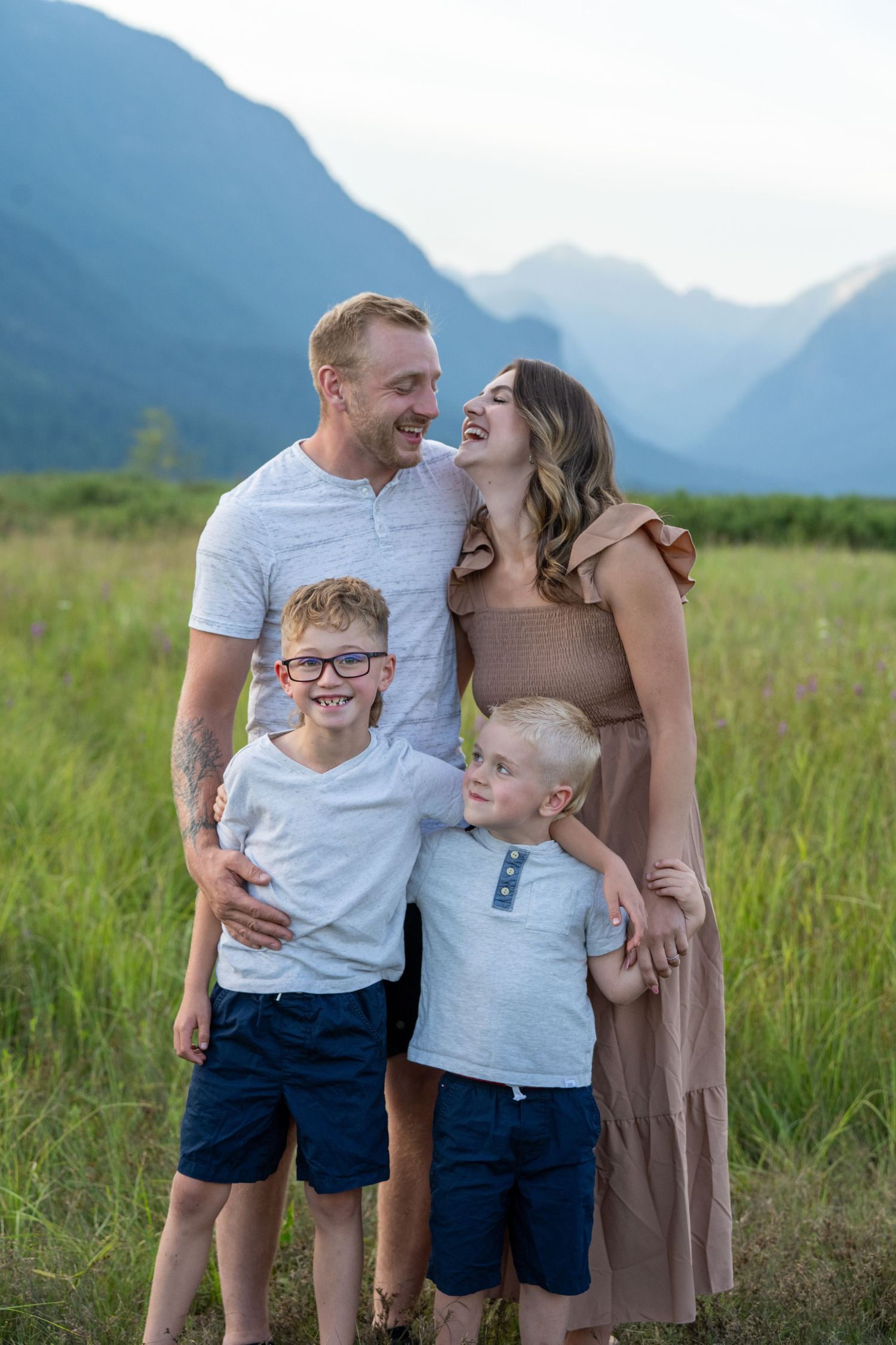 Happy family of four posing in a green meadow with mountain backdrop, parents laughing with two young boys.