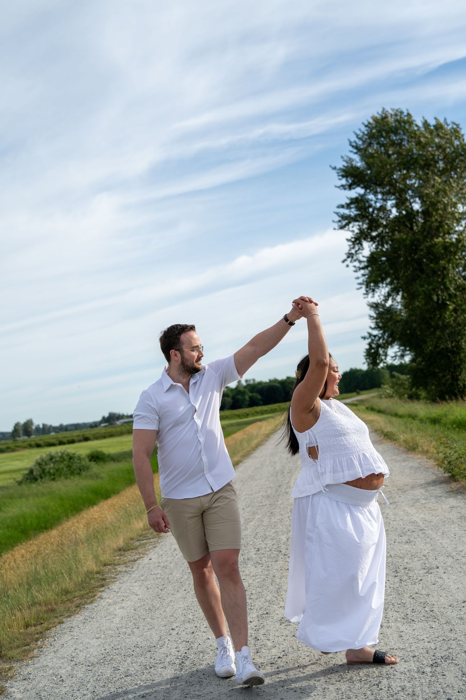Pregnant couple dancing on a rural gravel road, man in white shirt and shorts, woman in flowing white dress, green fields.