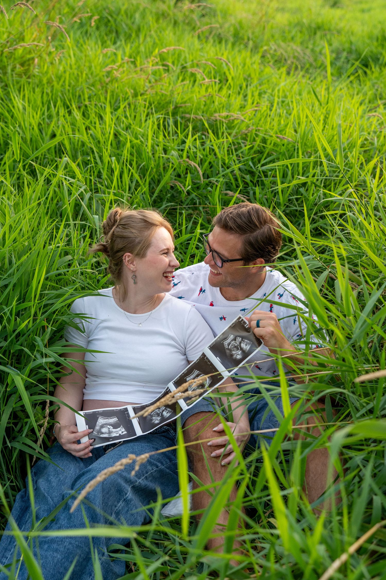 Happy couple sitting in lush green grass holding ultrasound photos, sharing a joyful moment during pregnancy announcement.