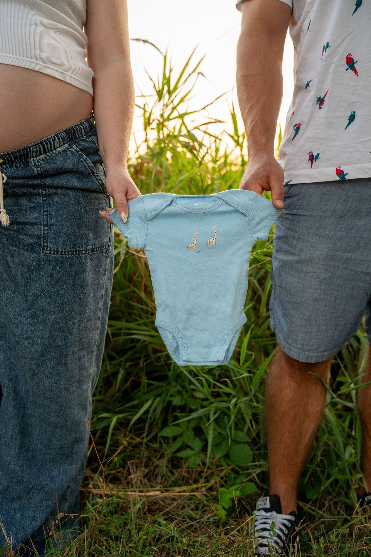 Couple holding a light blue baby onesie outdoors, with a pregnant woman's belly visible, announcing their pregnancy.