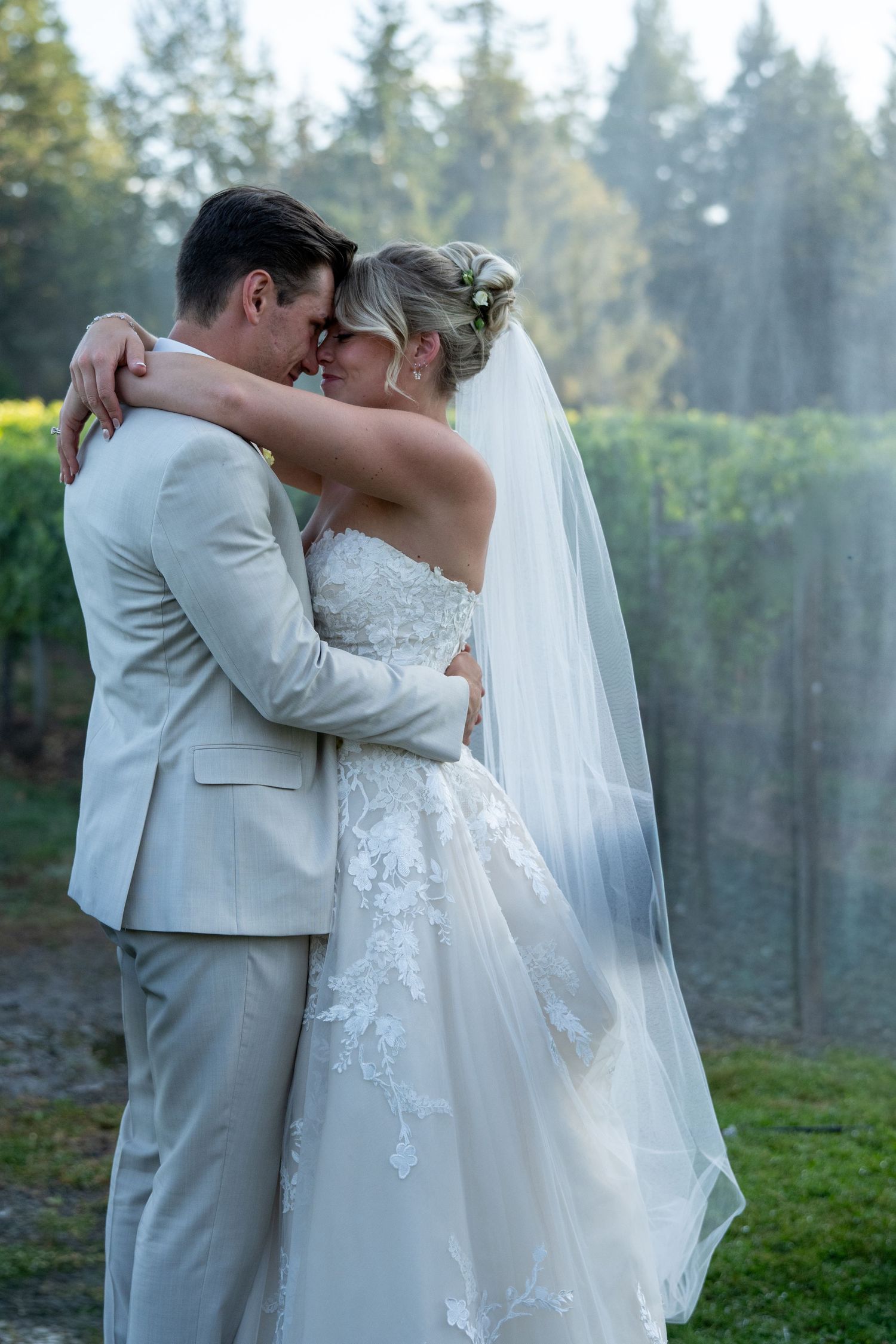 Bride and groom embrace tenderly in a lace wedding dress and tan suit, with a flowing veil and vineyard backdrop.