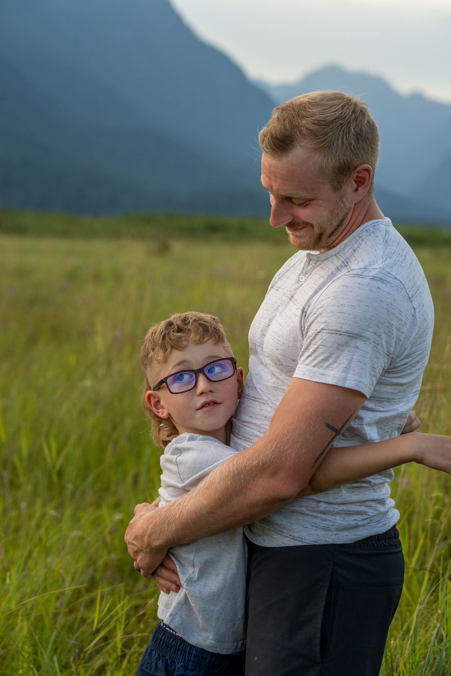A father smiles while hugging his young son wearing blue glasses in a green field with mountains behind them.