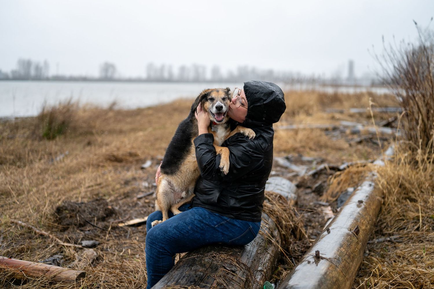 A person in winter clothing sits with a cat on a snowy landscape with mountains in the background. Port Coquitlam beach
