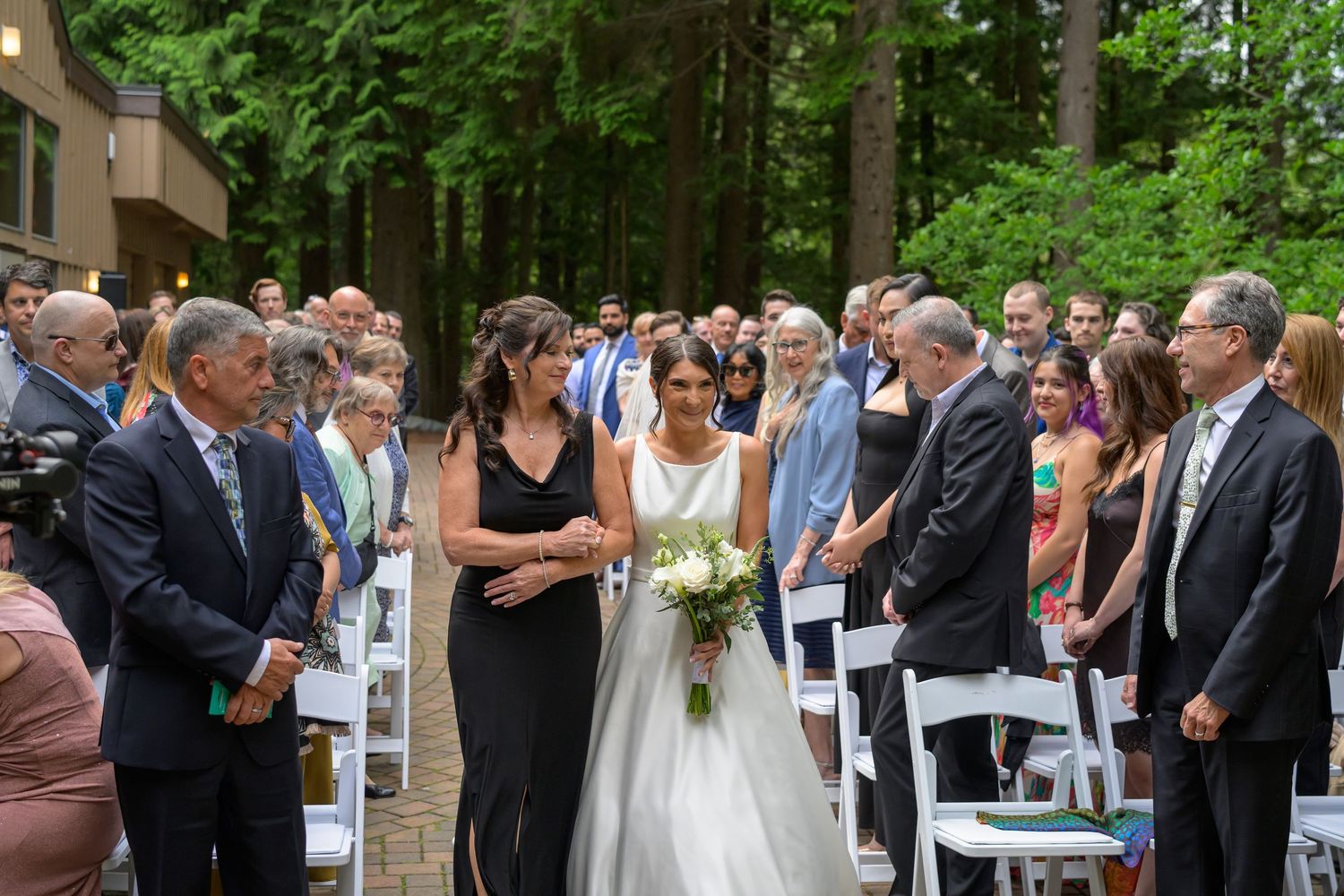 Bride walks down outdoor aisle in white gown holding bouquet, surrounded by standing wedding guests among tall trees.