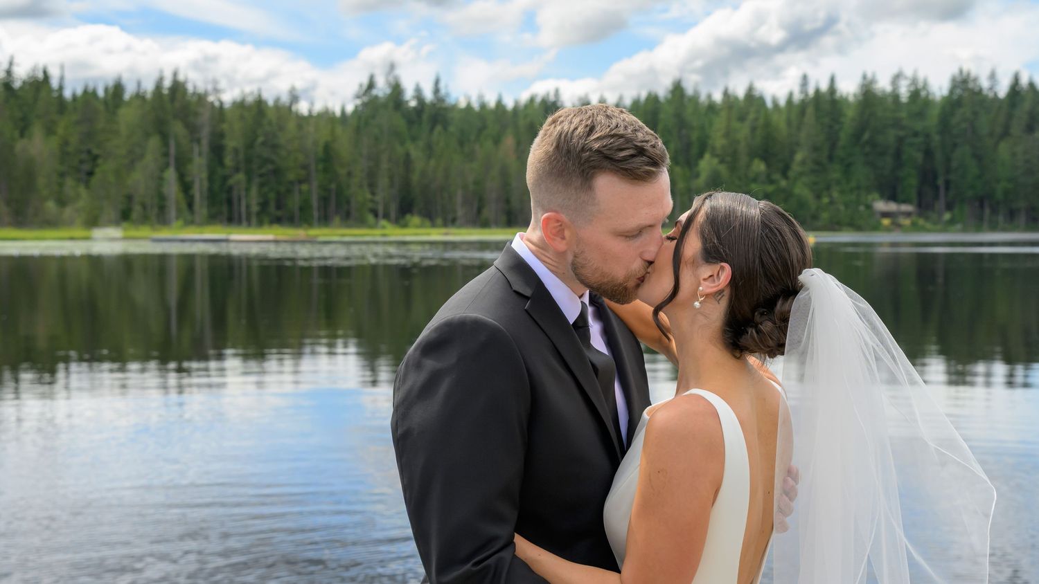 Bride and groom share a romantic kiss by a serene lake surrounded by lush green forest on their wedding day.