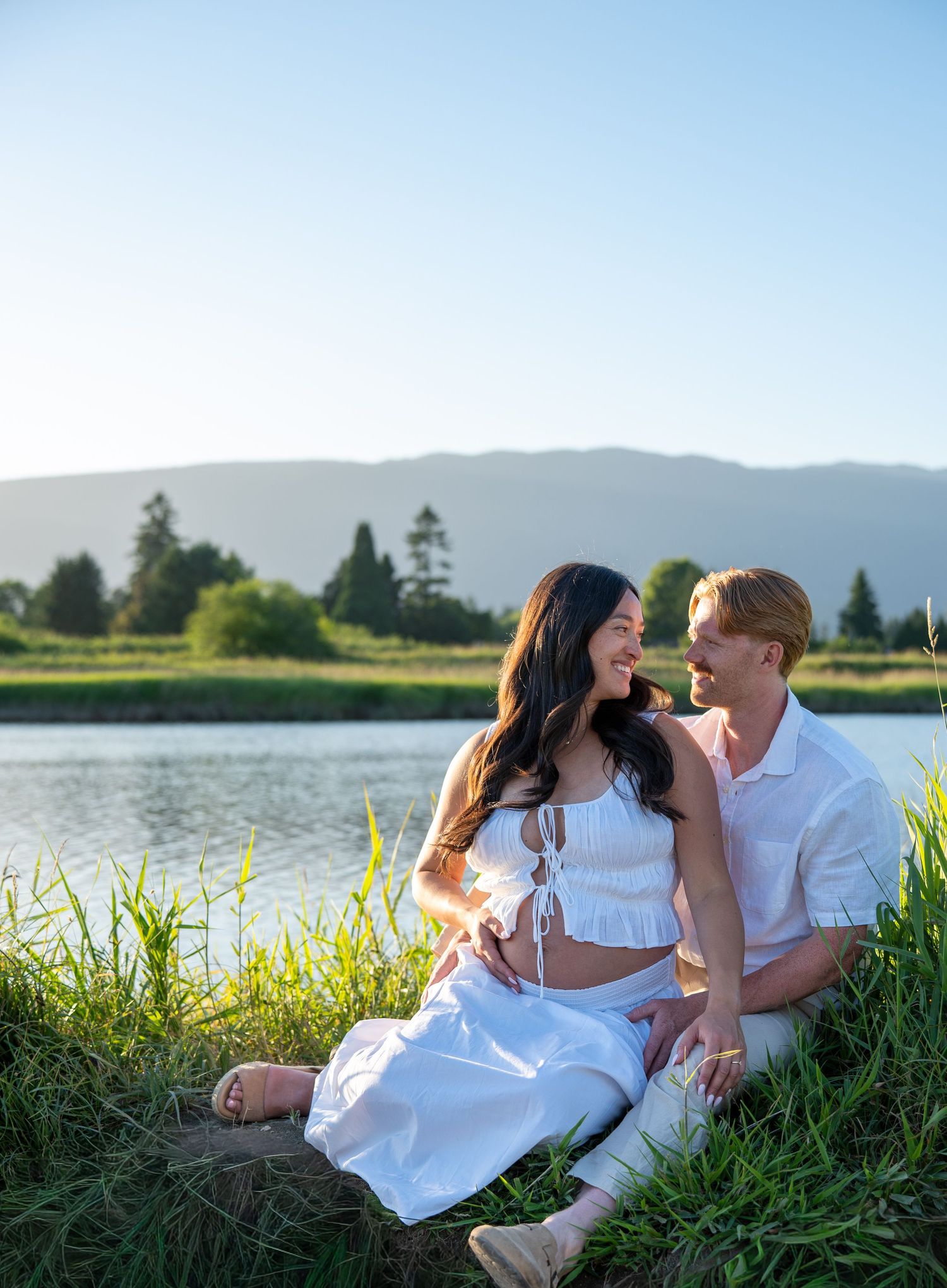 Couple in white outfits sitting together by a lake with mountain views on a sunny day.
