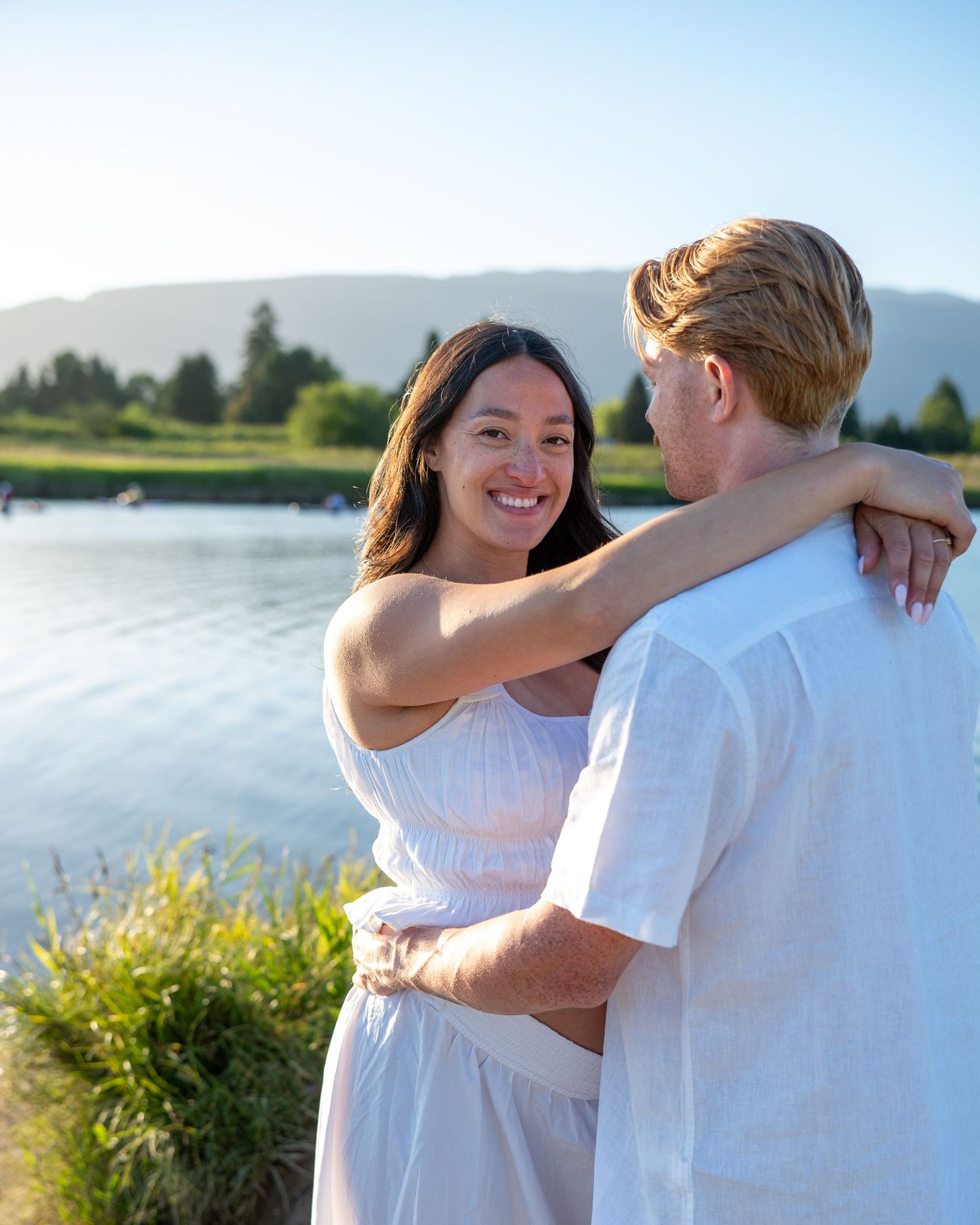 Smiling woman embracing a man near a scenic lake with mountains and greenery in the background.